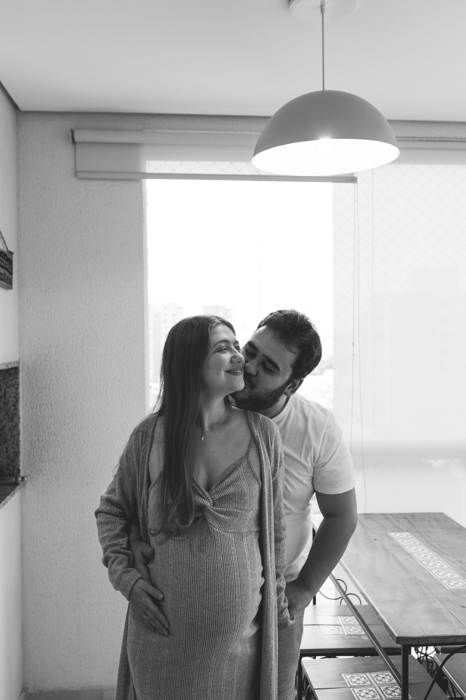 Black and white photo of man kissing pregnant partner near bright window indoors.