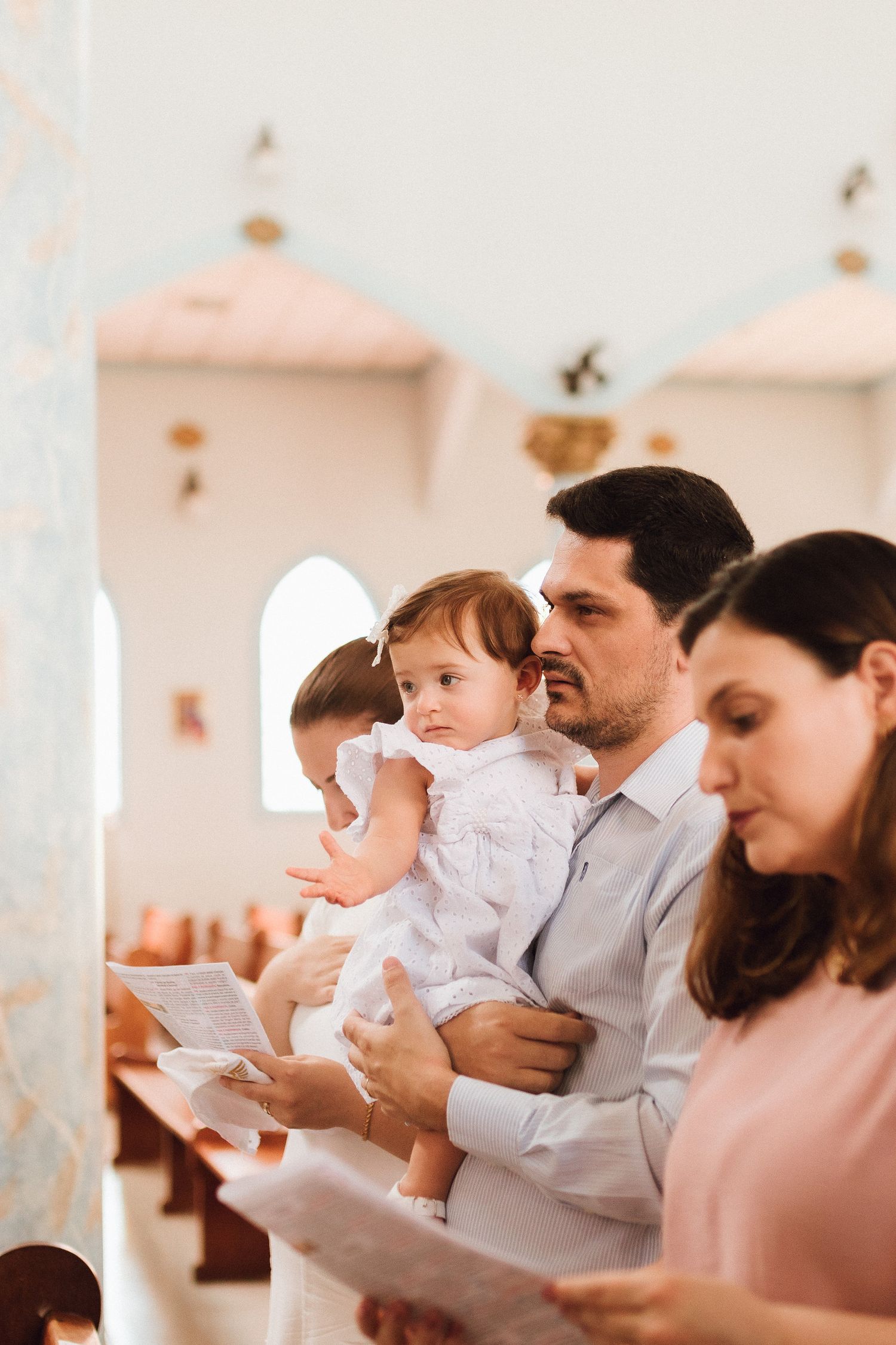 Family attending a church baptism ceremony, father holding baby girl in white dress while reading hymn sheets.