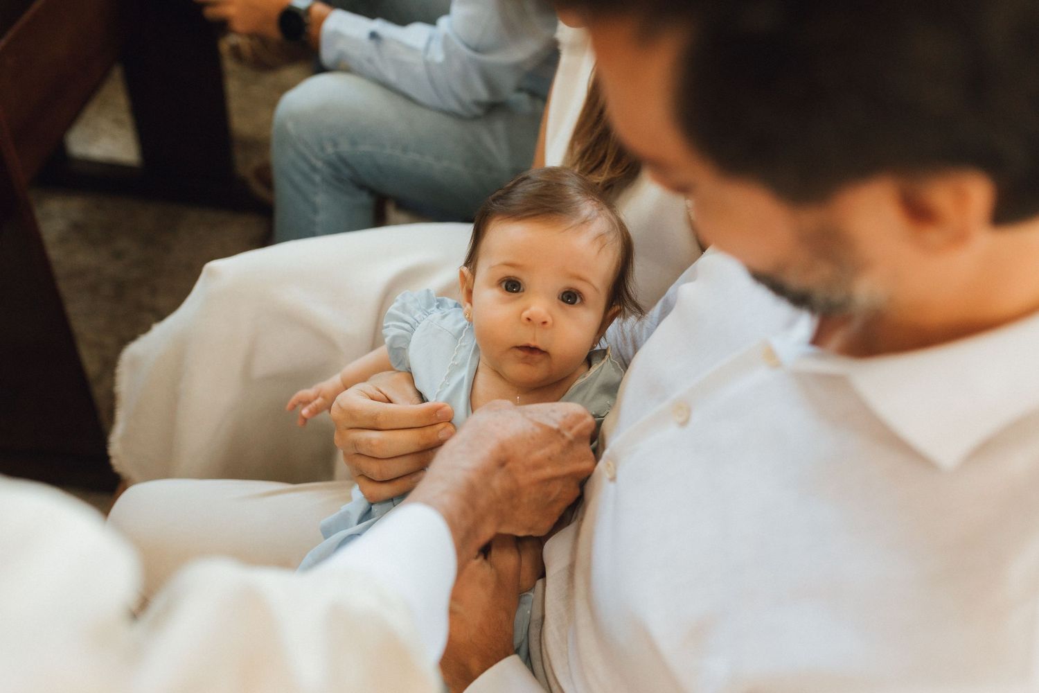 Adorable baby in blue outfit being held by an adult in a white shirt during a christening ceremony.