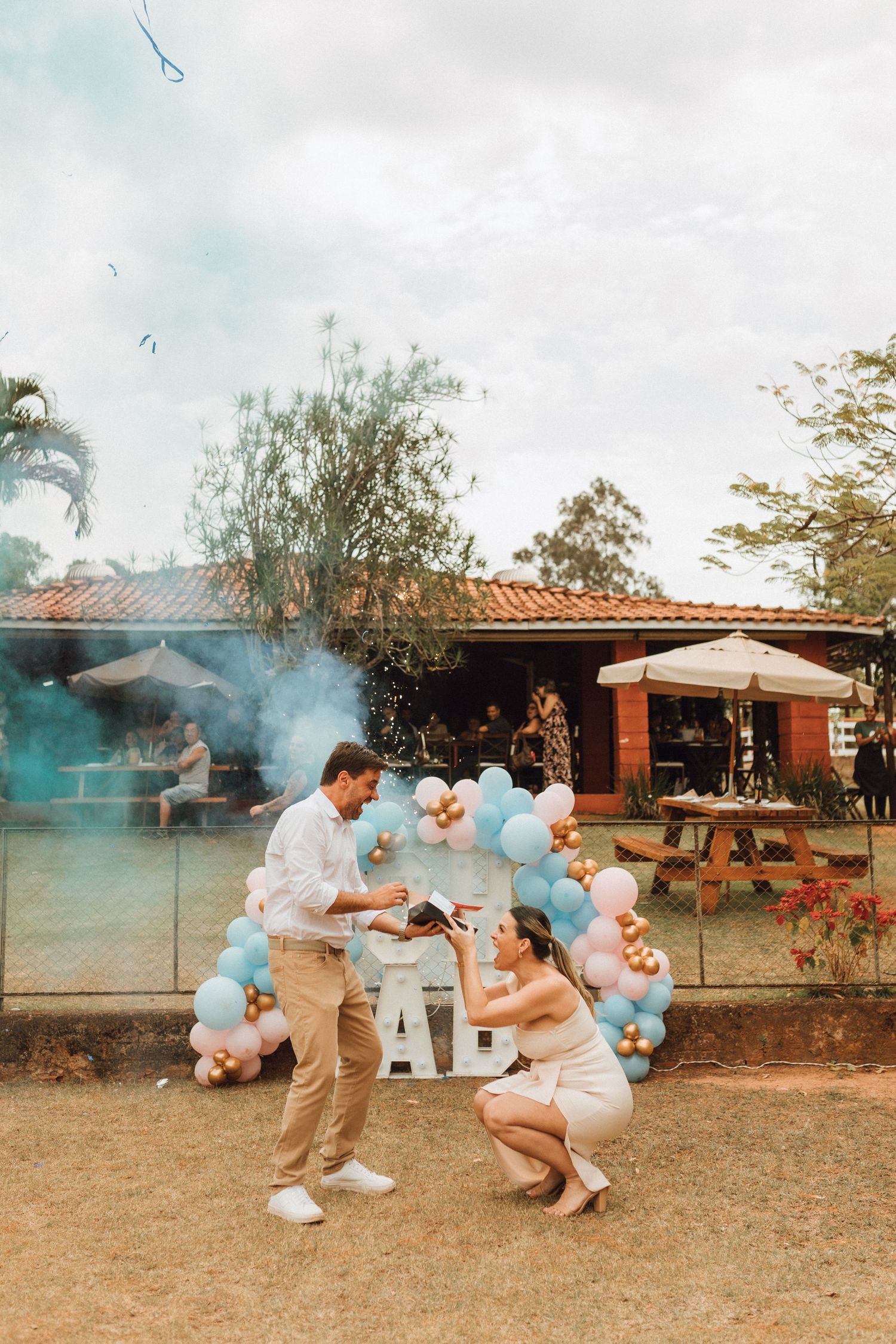 Couple crouching together during blue smoke gender reveal celebration outdoors with decorative balloon arch and OH BABY sign.