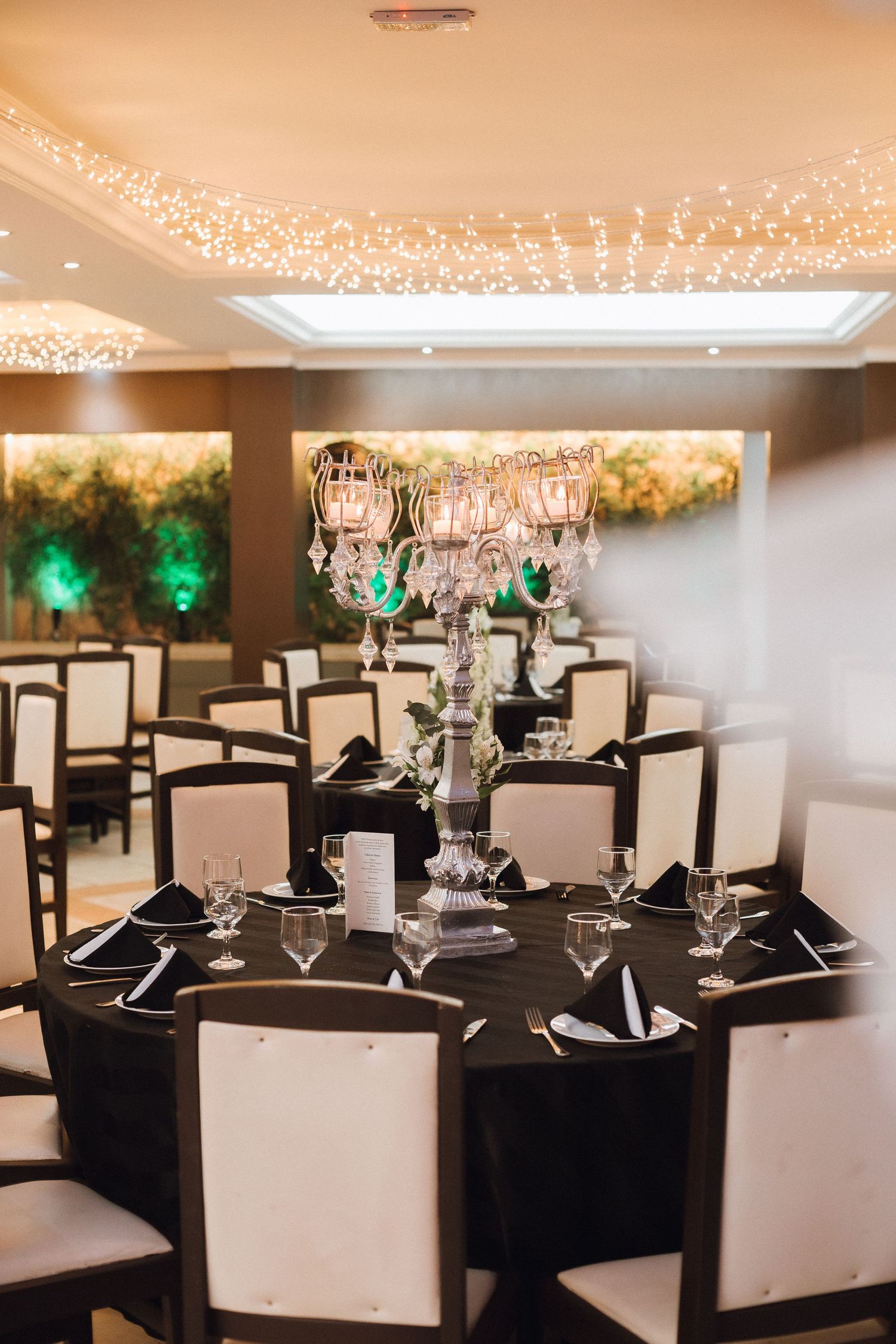 Elegant banquet hall with black-draped round tables, white chairs, and crystal candelabras under string lights.