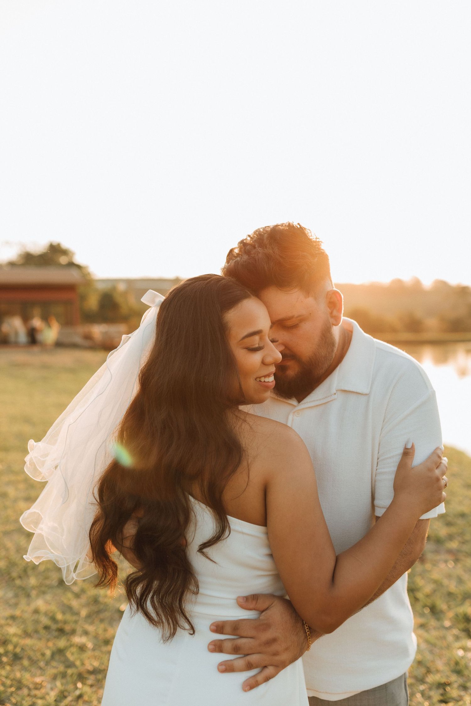 Bride and groom embracing at golden hour sunset outdoors, bride wearing white dress and veil, smiling joyfully.