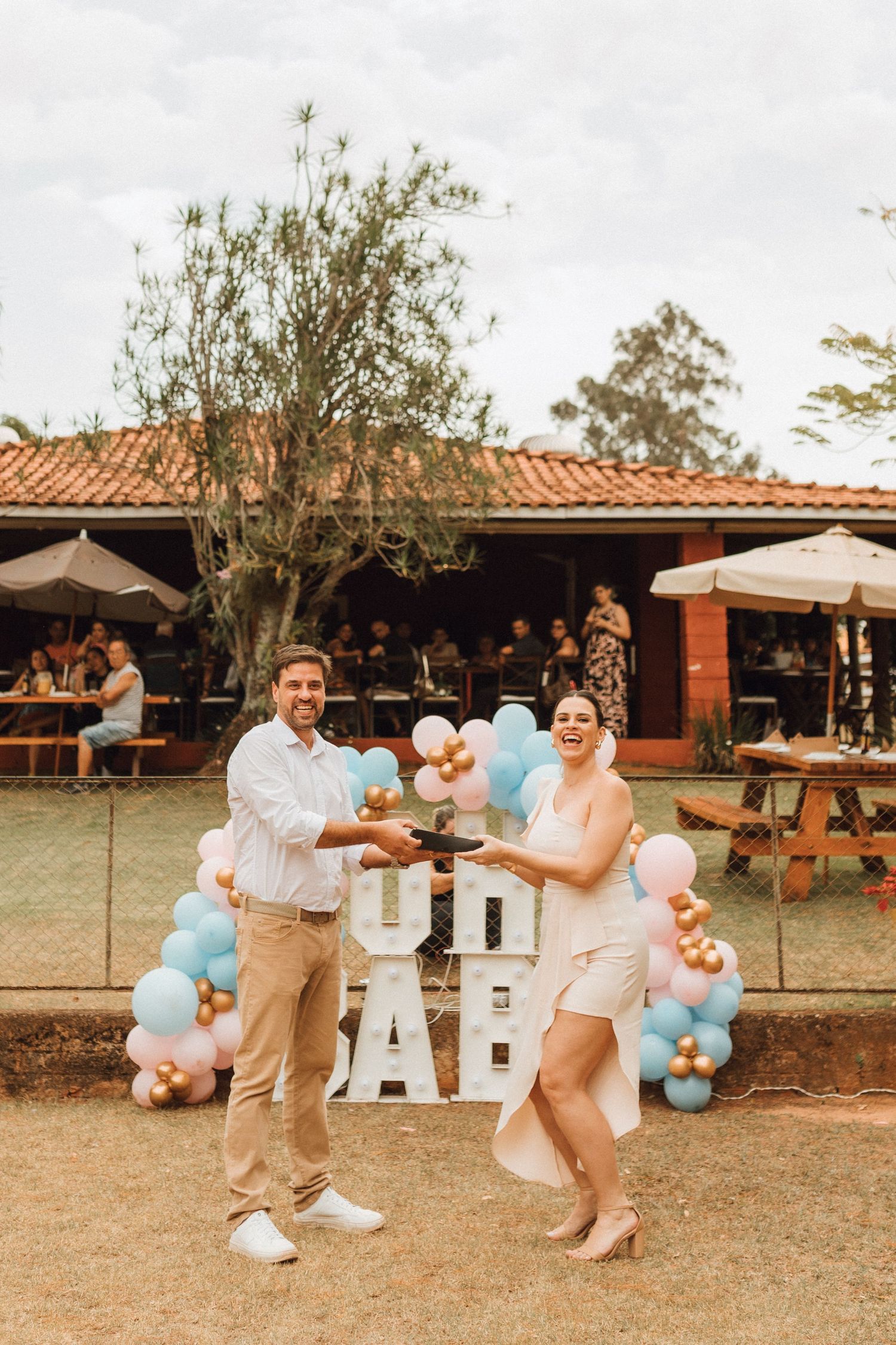 Couple laughing and holding a cake at a gender reveal party with pink and blue balloon decorations outdoors.