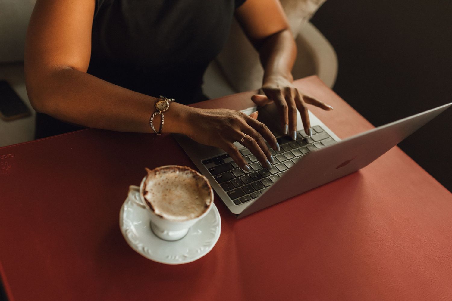 Woman typing on laptop at red table with cappuccino coffee cup, wearing gold bracelet bracelet.