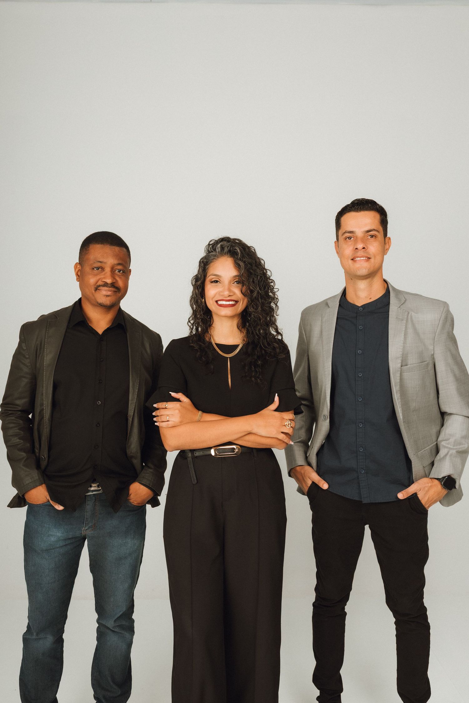 Three diverse professionals in smart casual attire posing together against a light gray studio background.
