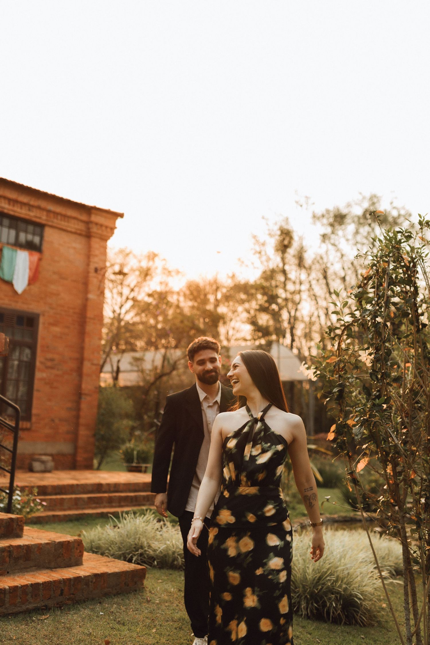 Couple walking in garden at golden hour, woman in floral dress smiling at bearded man in dark suit near brick building.