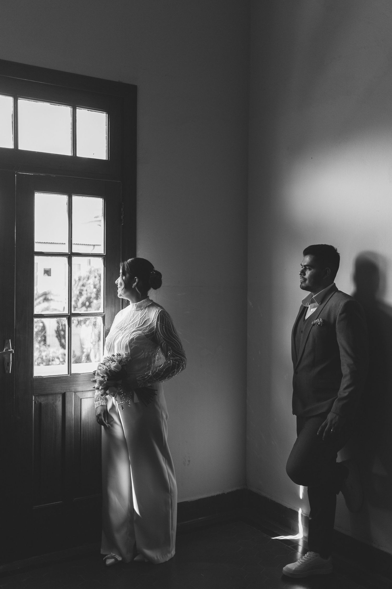 Black and white wedding photo of bride holding bouquet by window while groom stands nearby in elegant indoor setting.