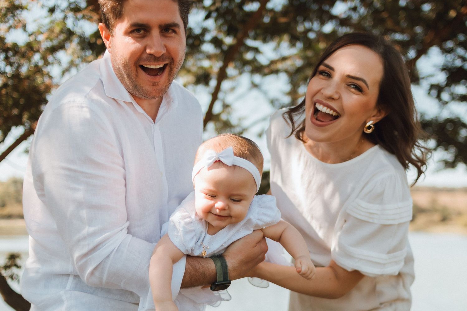Happy family with laughing parents holding their smiling baby outdoors in a bright, sunny setting.