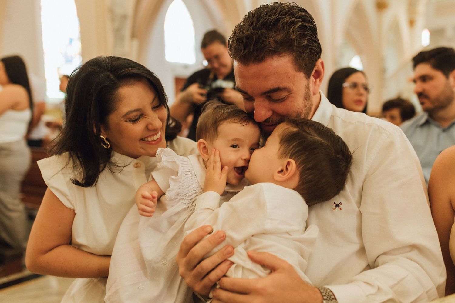 Family in white outfits sharing a joyful moment at a church baptism ceremony, parents lovingly embracing two babies.