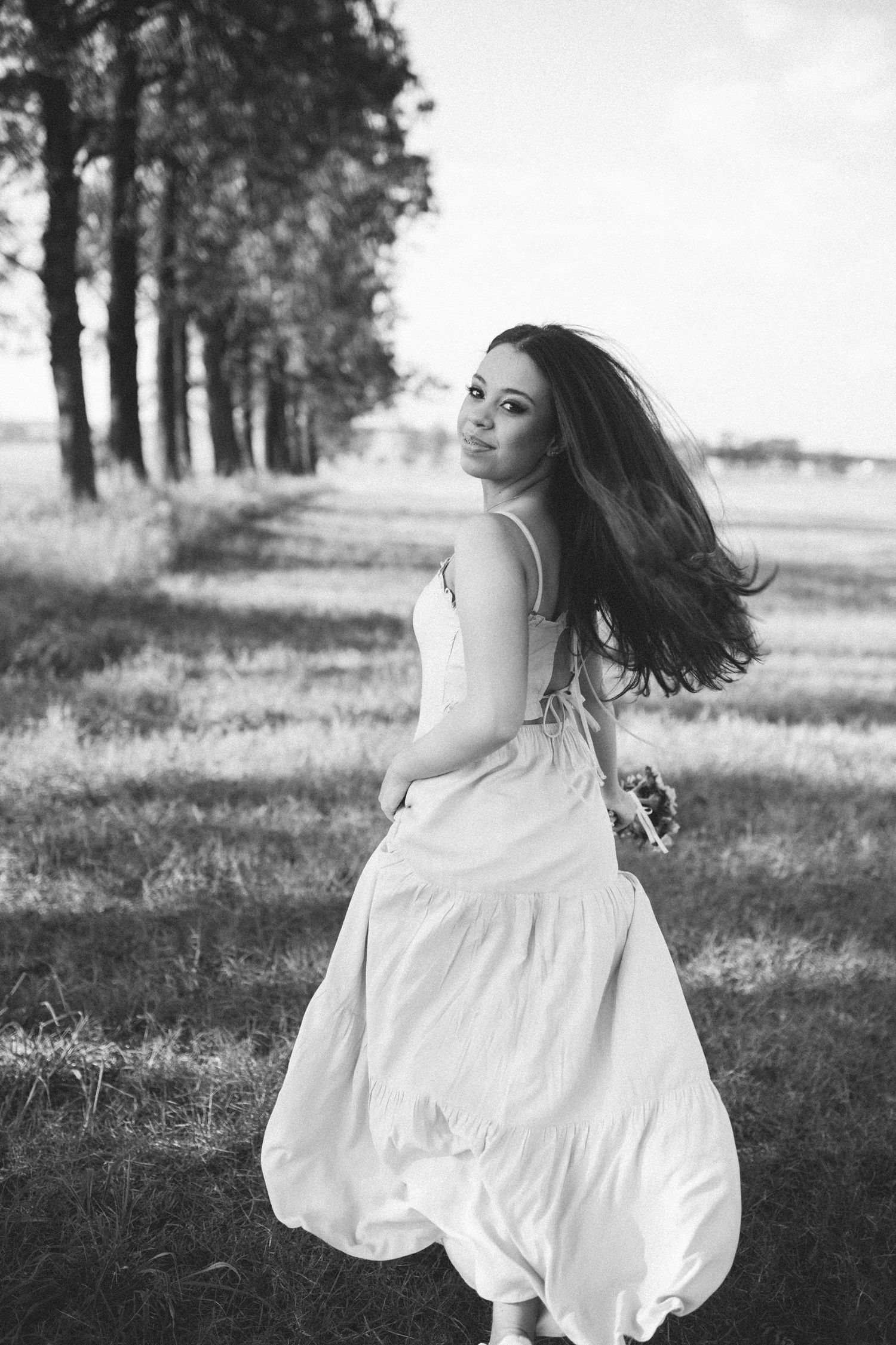Black and white photo of a woman in a flowing white dress spinning outdoors near trees on a sunny day.