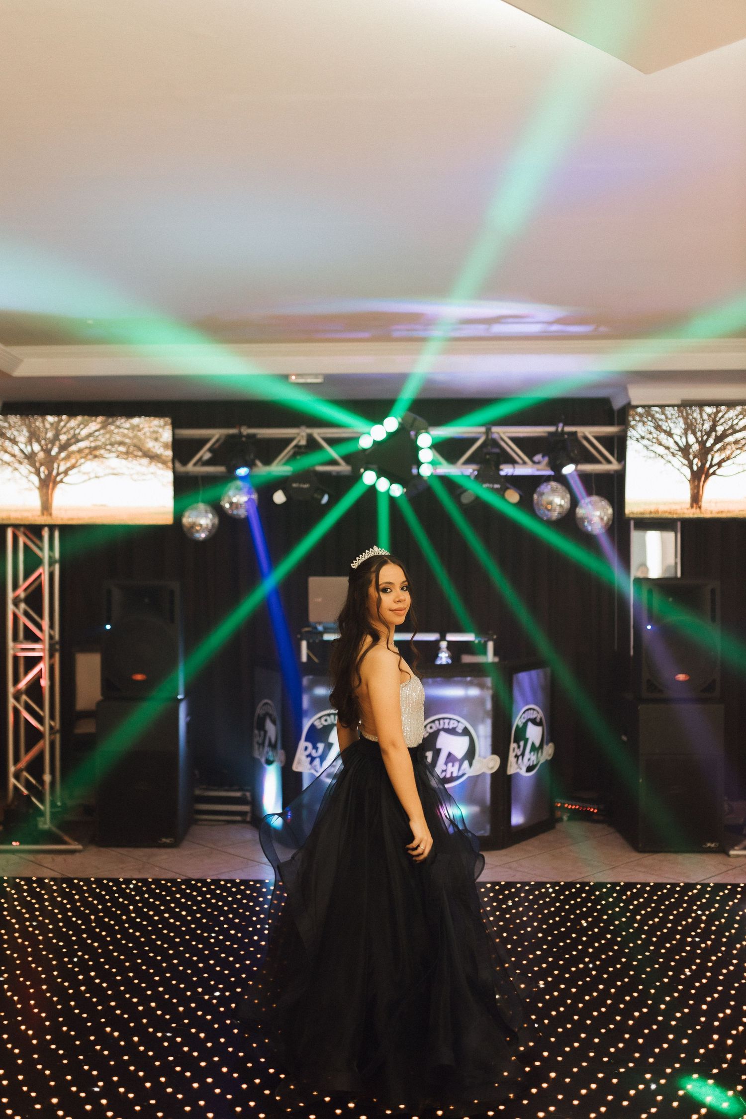 Quinceañera girl in black ballgown and tiara poses on LED dance floor with green laser lights.