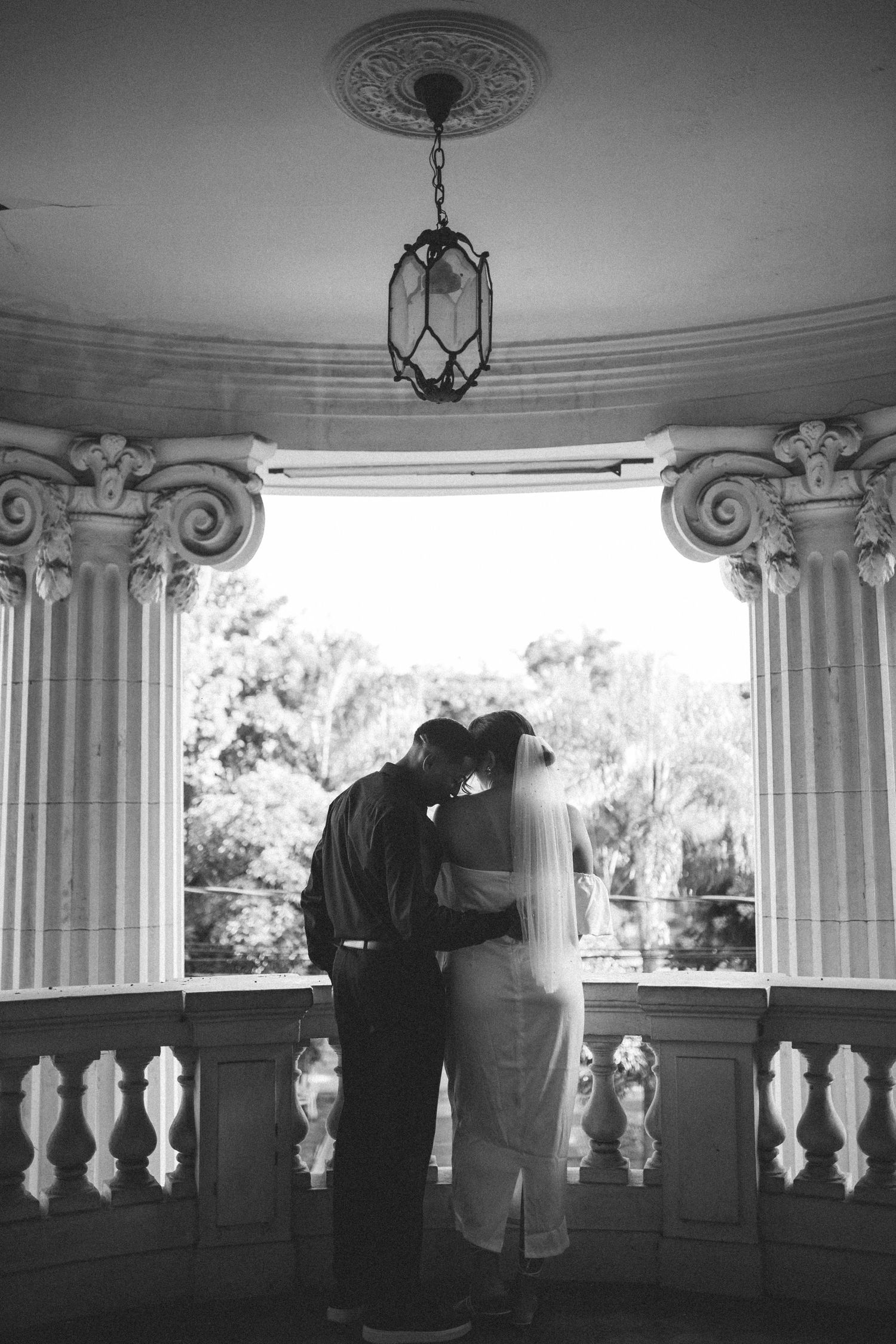 Black and white silhouette of bride and groom embracing on elegant columned balcony with hanging lantern.