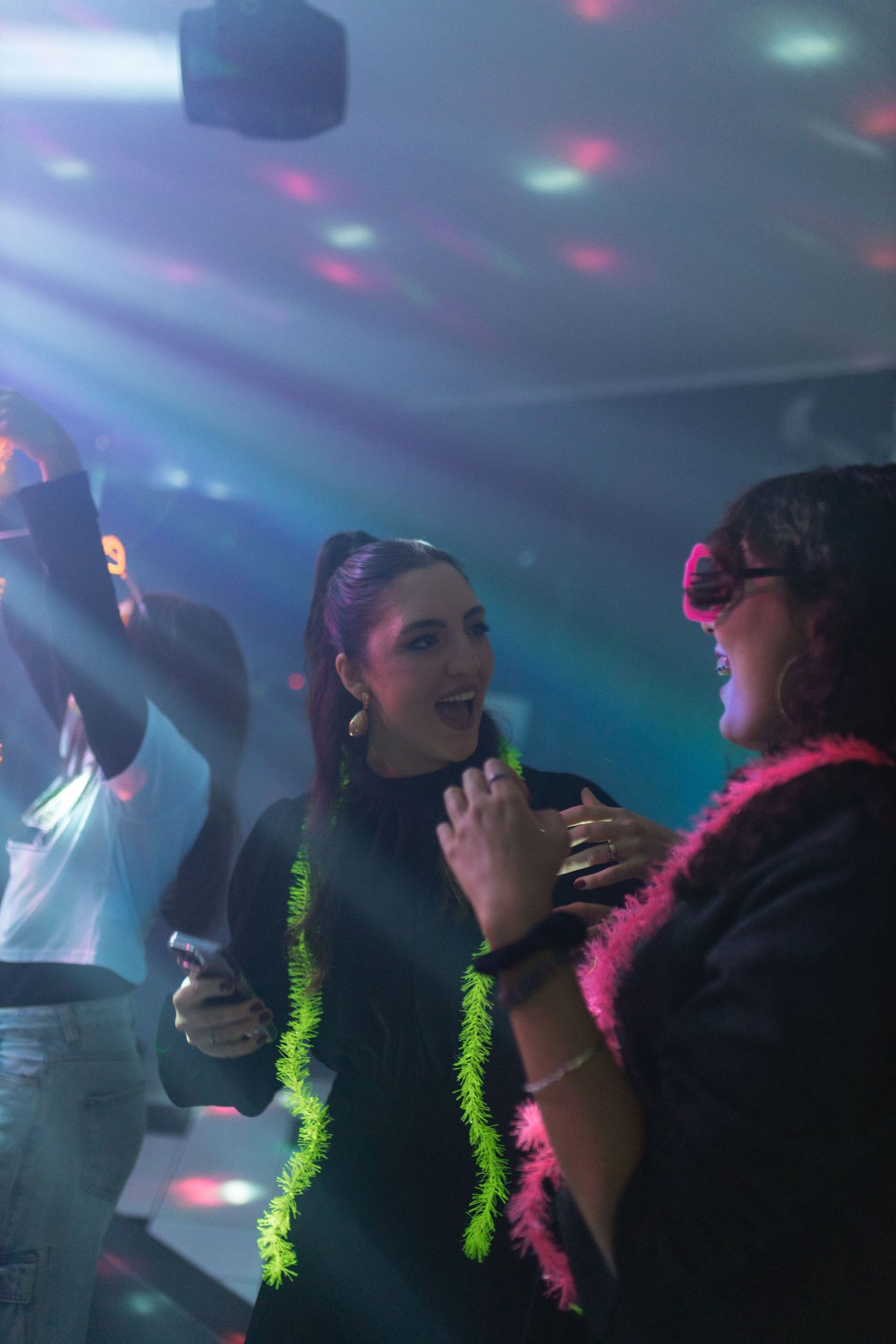 Women dancing joyfully at nightclub party under colorful disco lights, wearing festive neon accessories.