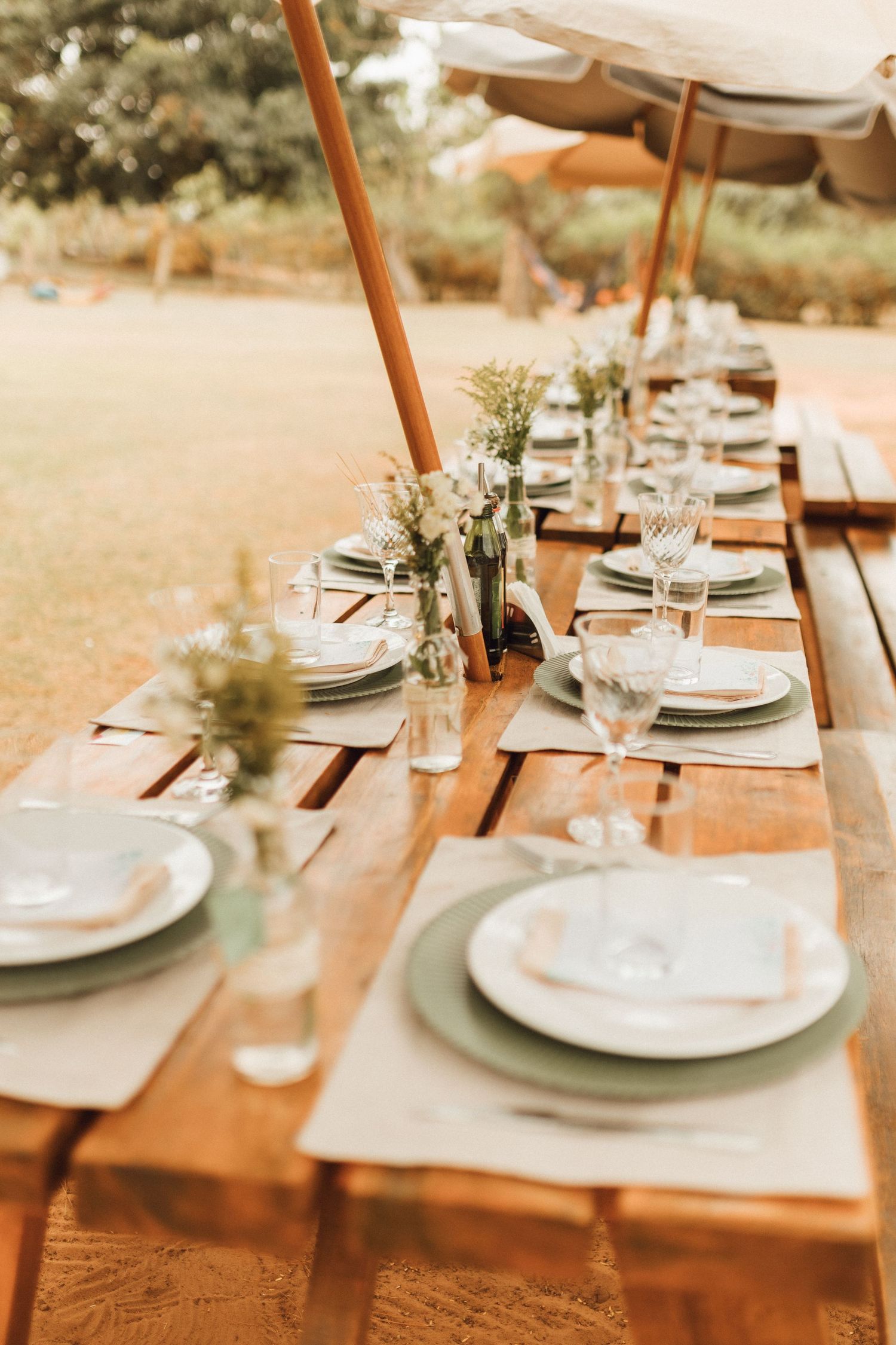 Rustic outdoor wedding reception table set with white plates, wine glasses, and floral centerpieces on a wooden farm table.