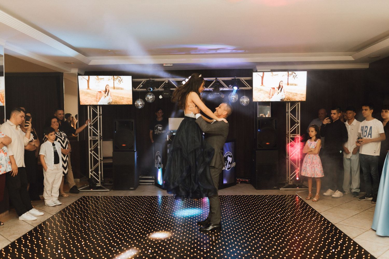 Couple shares romantic lift dance on star-lit dance floor surrounded by guests at festive celebration.