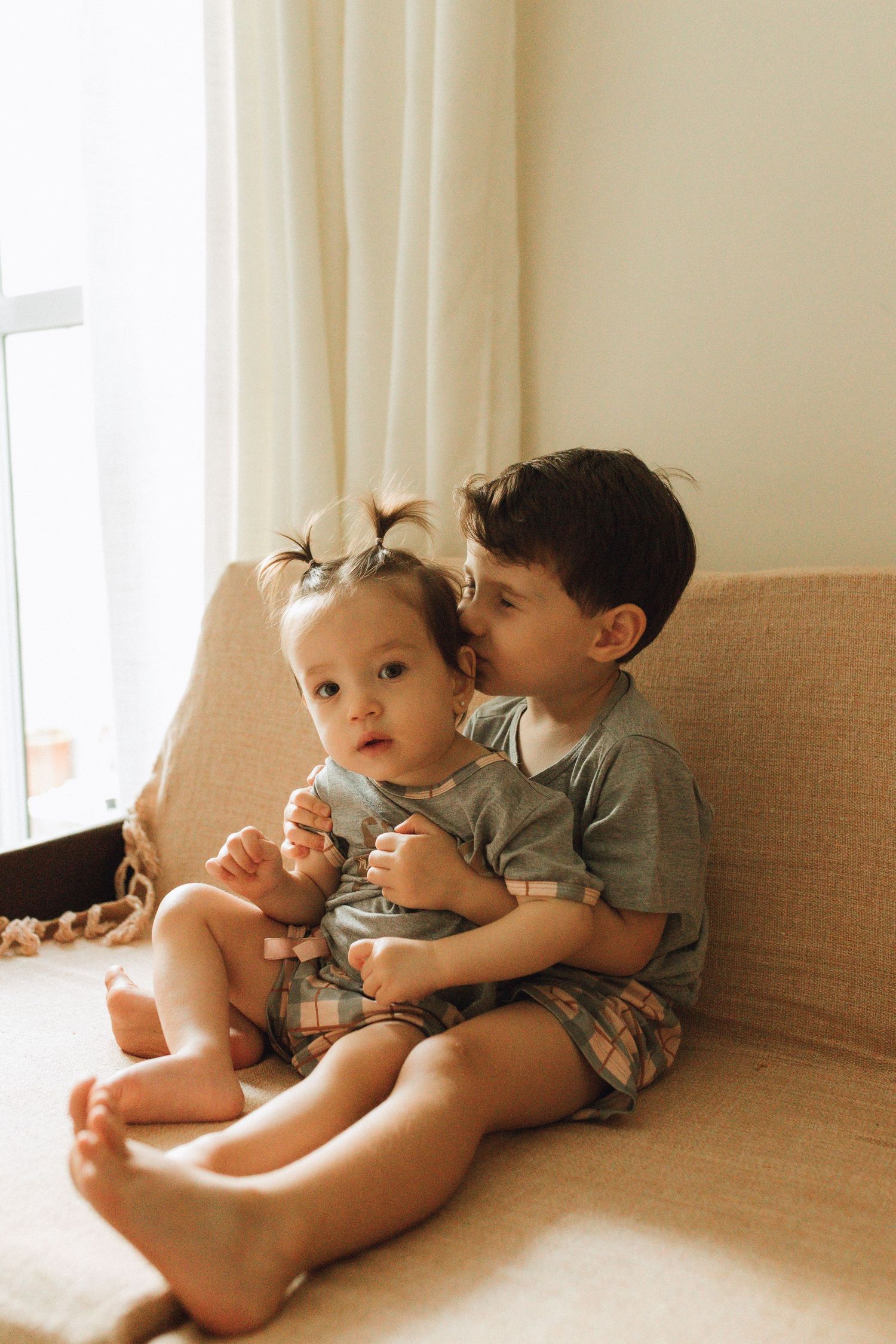 Two young siblings sitting together on a beige sofa, older boy hugging baby girl near a bright window.