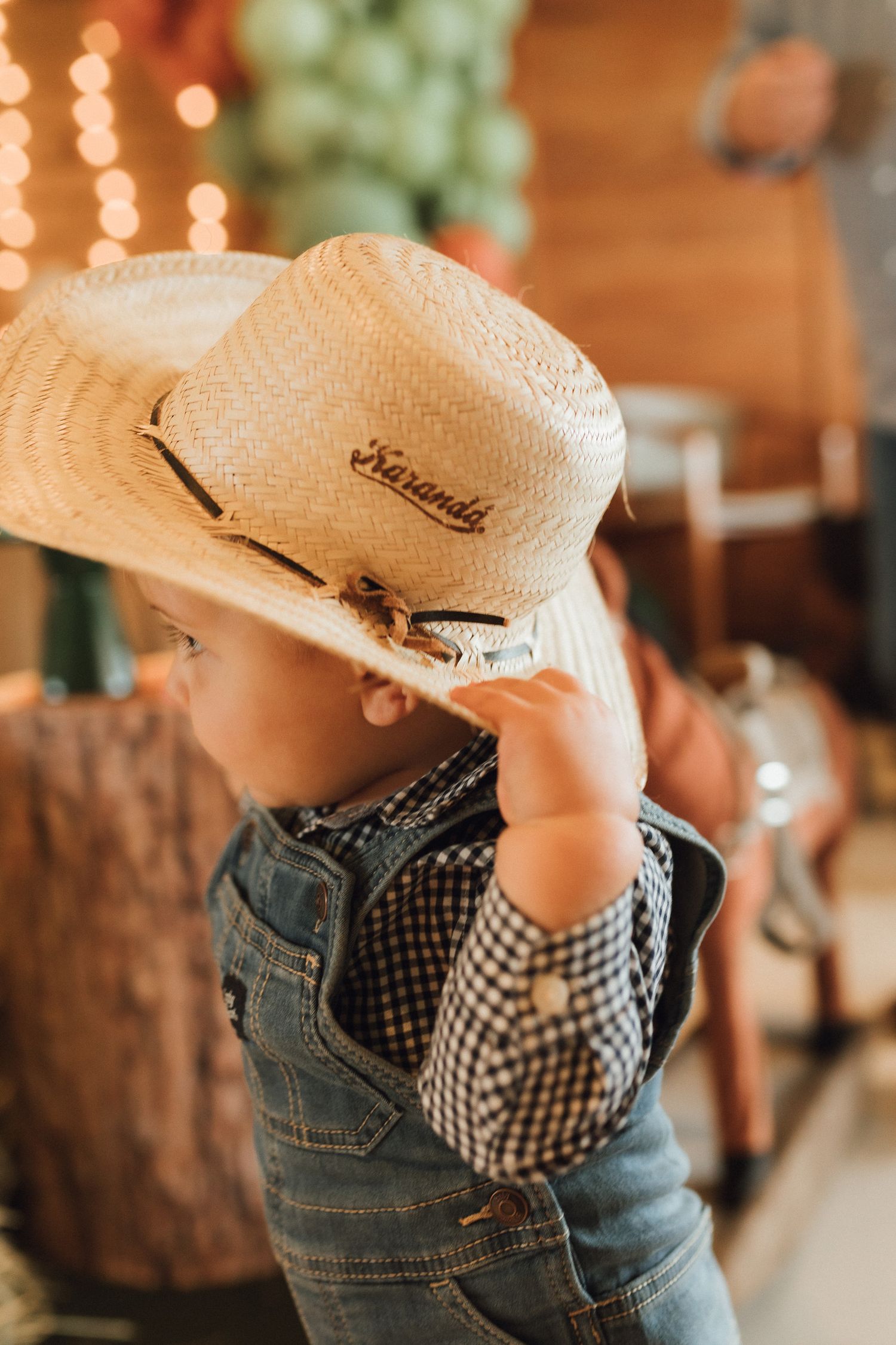 Toddler in denim overalls and plaid shirt wearing a straw Karanda cowboy hat at a rustic western party.