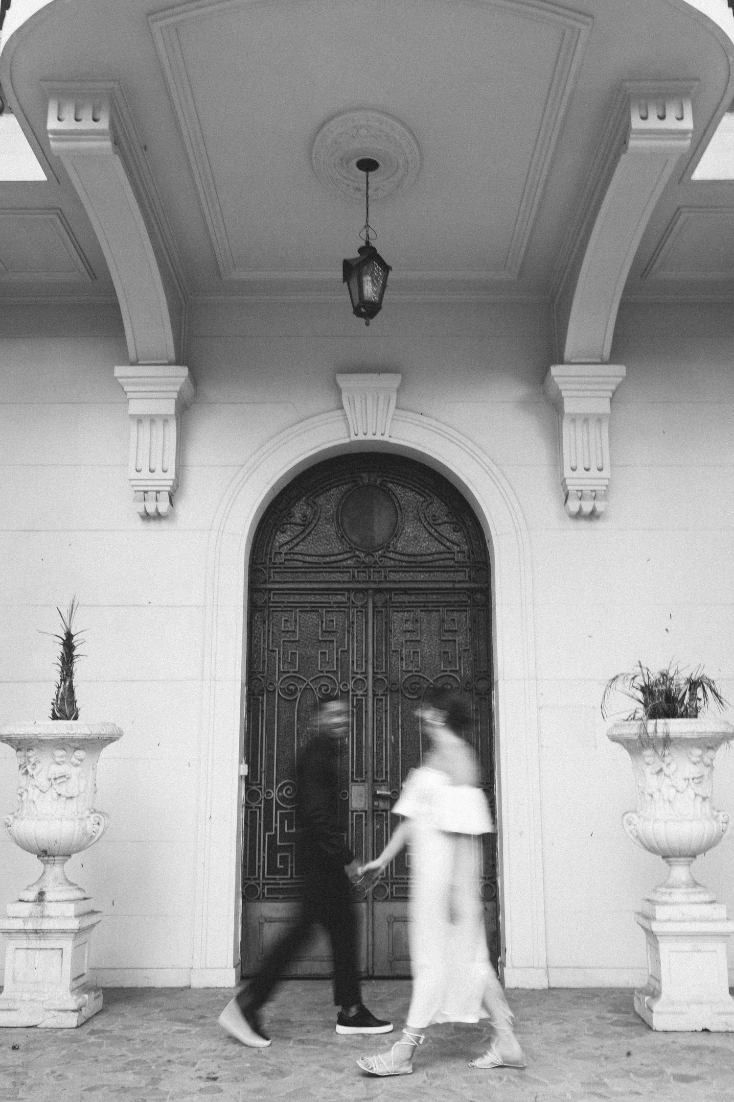 Blurred couple holding hands walking past ornate arched doorway with decorative urns in black and white.