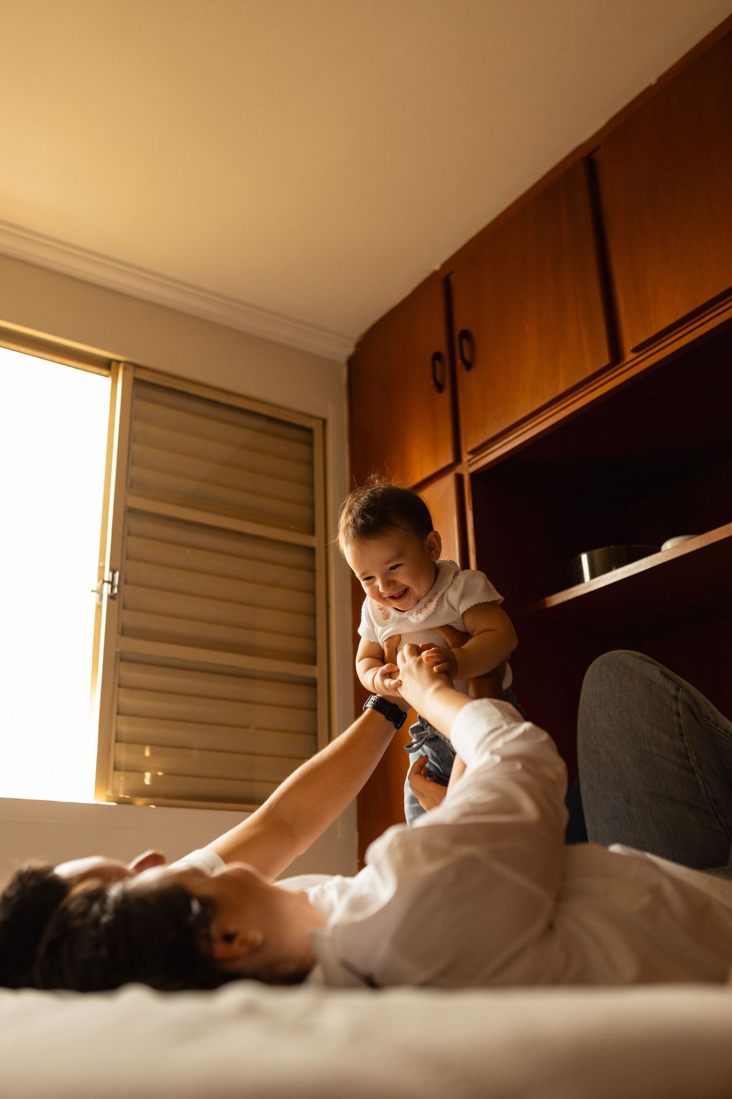 Parent lying on bed lifting smiling baby in the air in a warmly lit bedroom.