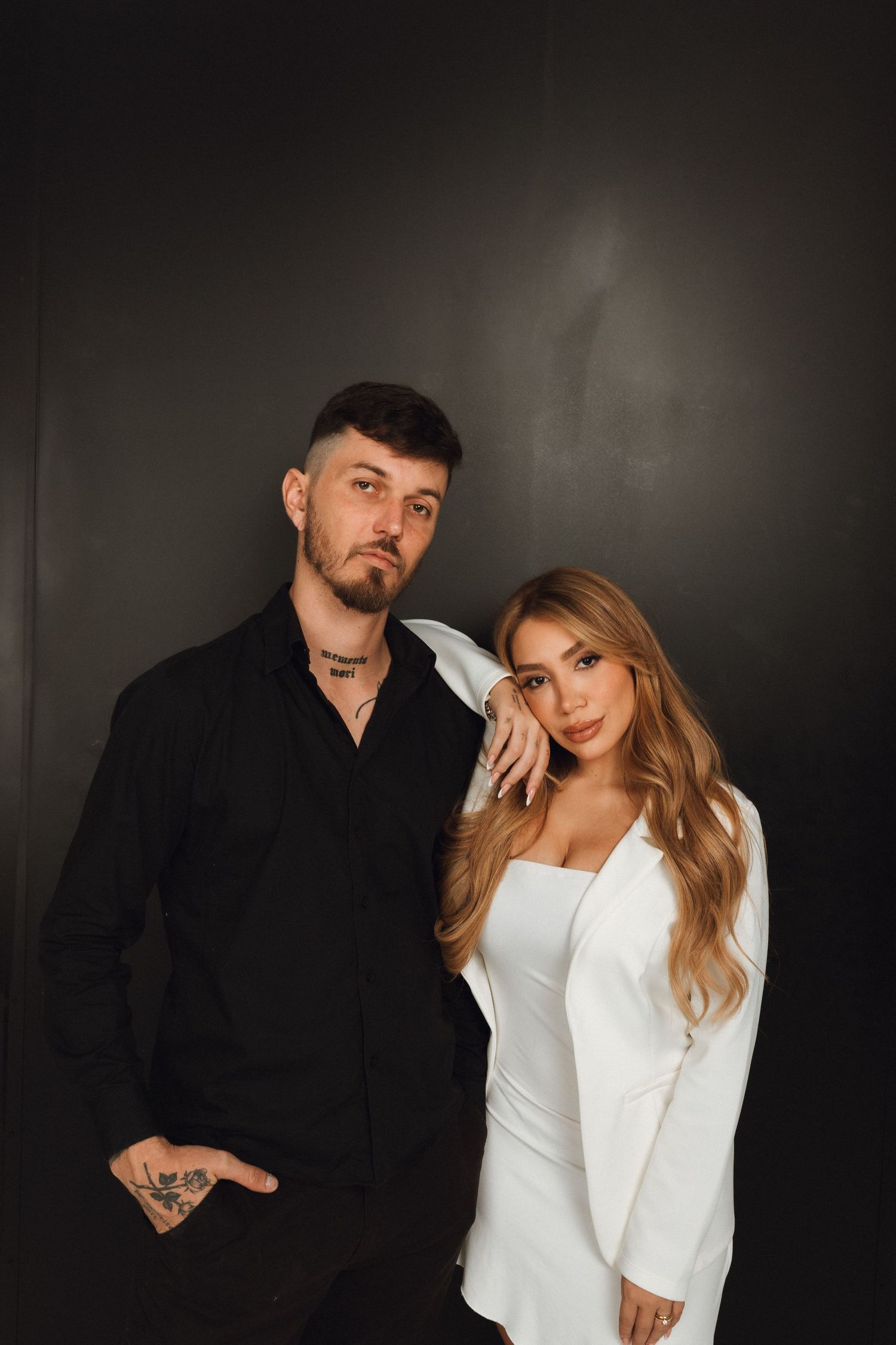A tattooed man in black shirt poses with a blonde woman in white blazer dress against a dark background.