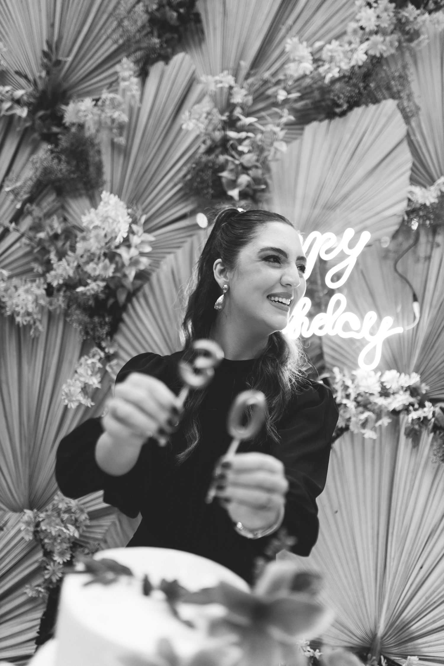 Black and white photo of smiling woman cutting birthday cake in front of floral backdrop with Happy Birthday neon sign.