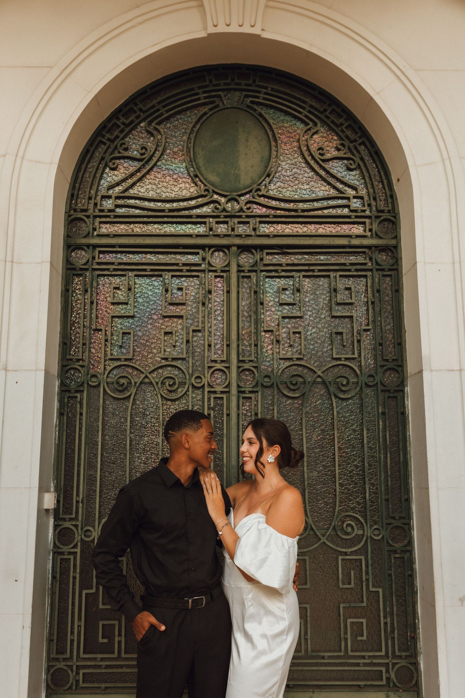Couple in wedding attire smiling at each other in front of ornate green arched door.