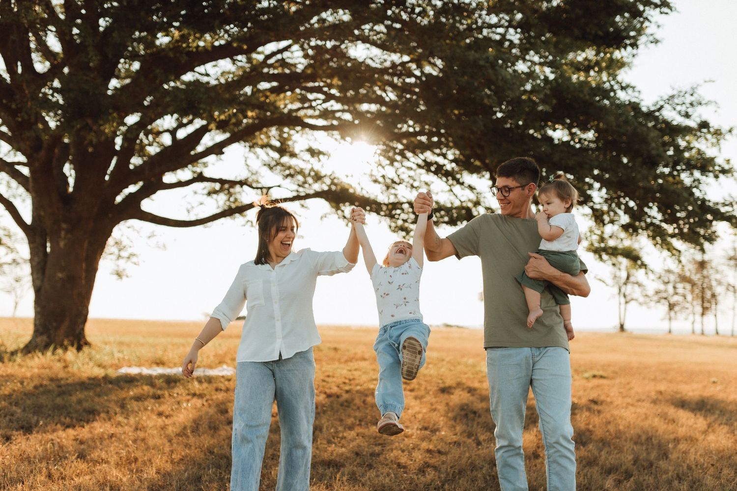 Happy family swinging toddler between parents under a large tree in a sunny field.