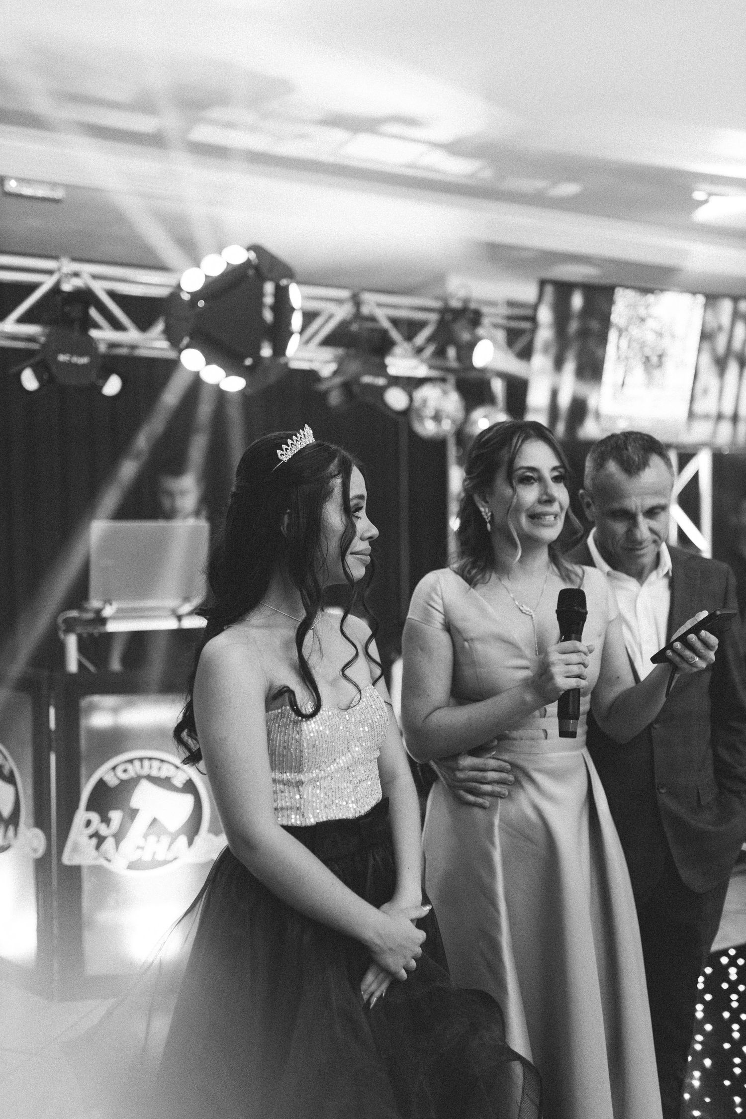 Black and white photo of quinceañera girl in tiara with woman speaking into microphone at party.