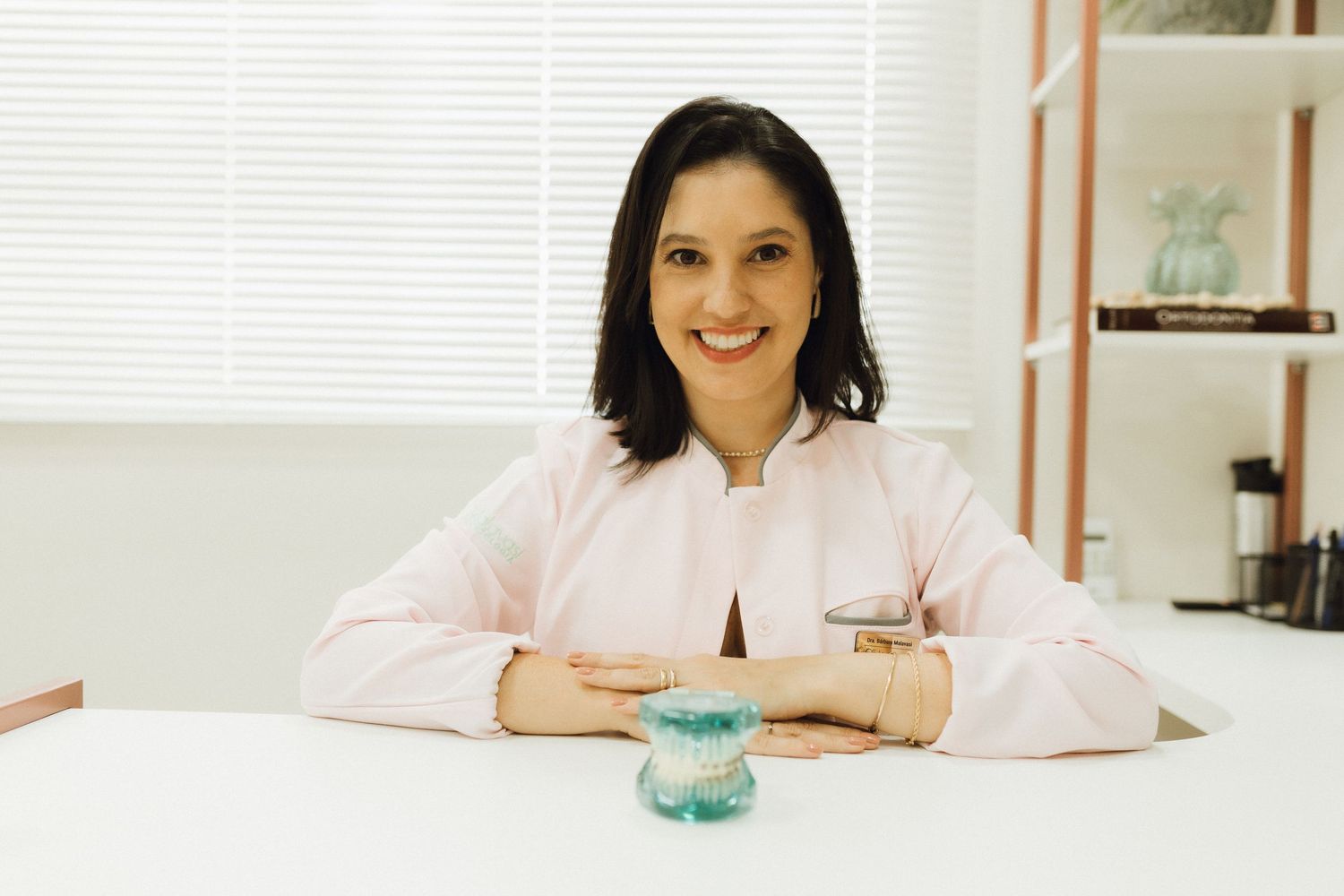 Smiling woman in pink blouse sitting at white desk with folded arms, bright modern office background.