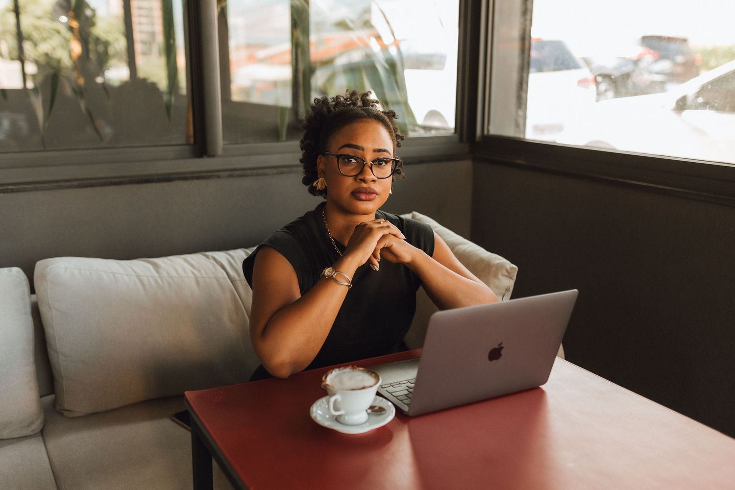 A young Black woman with glasses sits at a cafe table with a laptop and coffee cup, looking thoughtfully at the camera.
