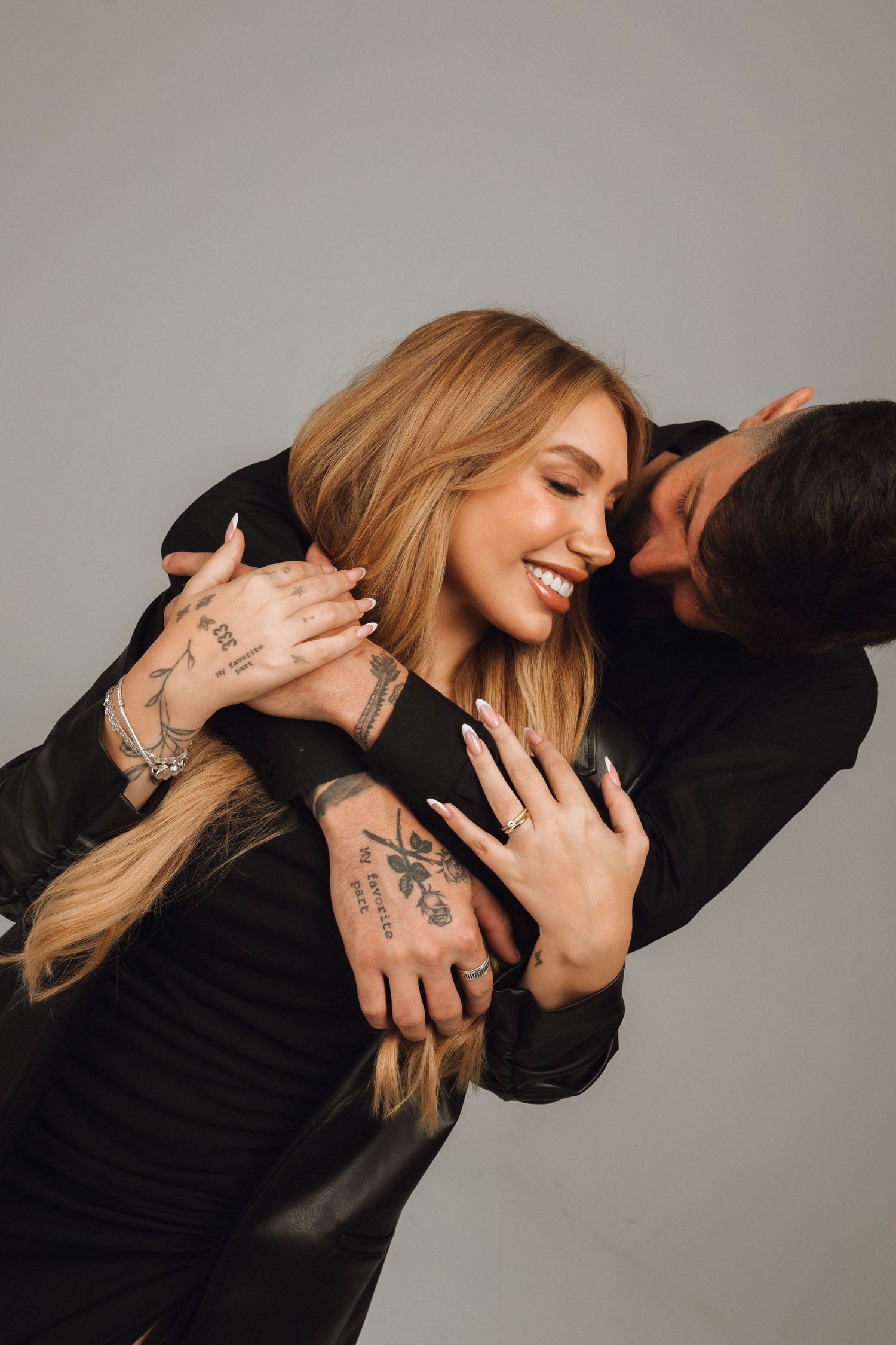 Smiling blonde woman with tattoos embraced from behind by a man, both wearing black outfits against a gray background.