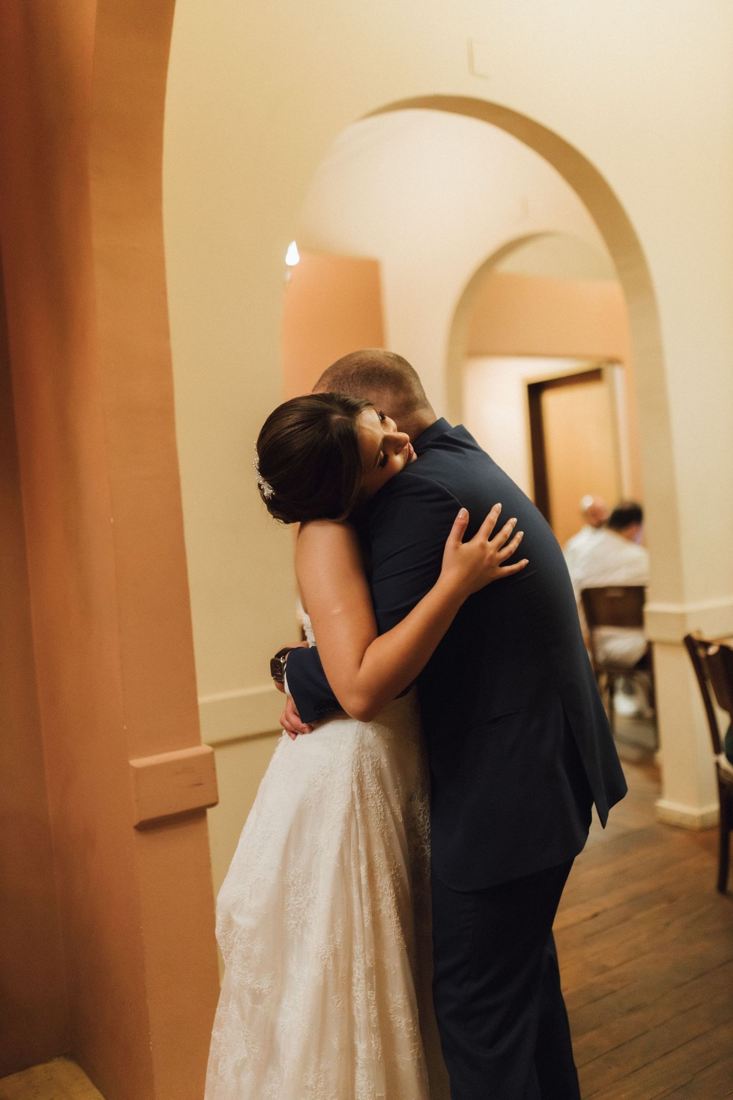 Bride and groom share a tender hug in a warmly lit venue with elegant arched doorways.