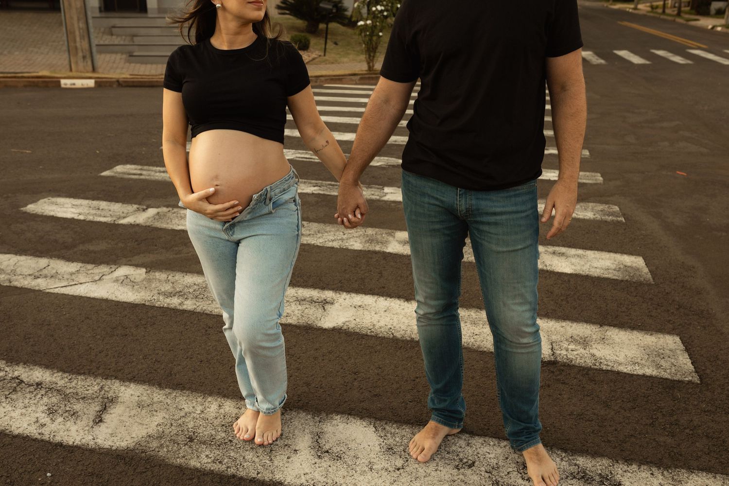 Pregnant couple holding hands barefoot on a zebra crossing, woman cradling baby bump in casual jeans.