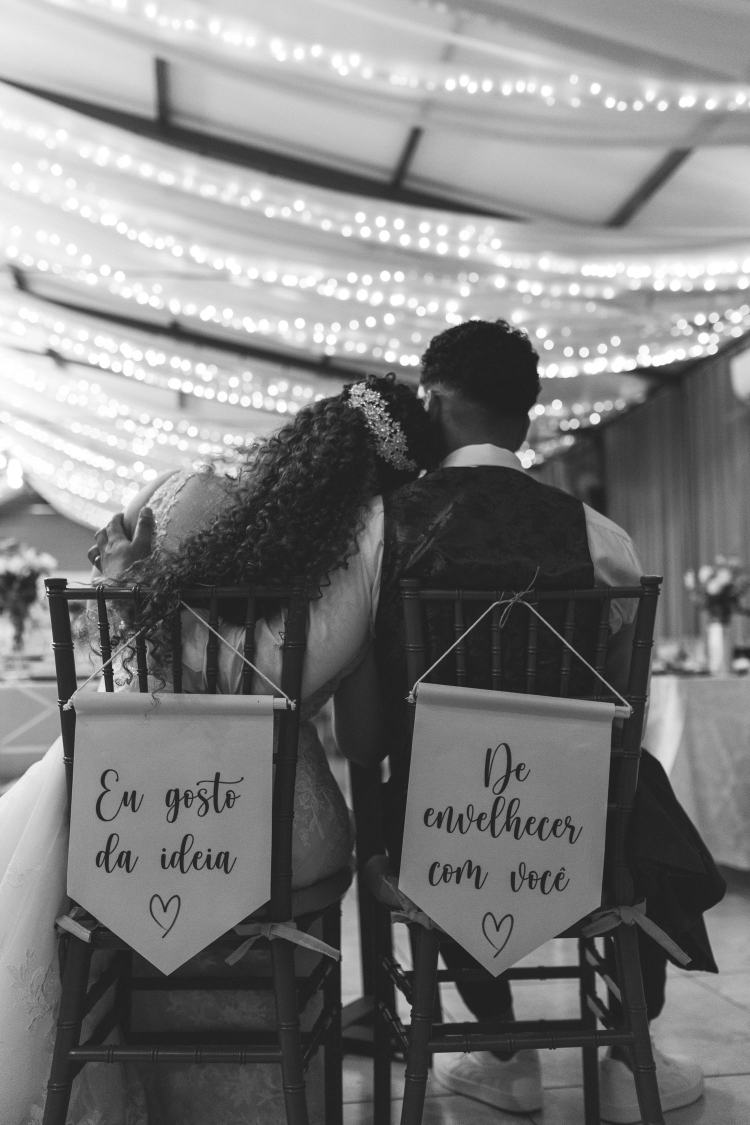 Black and white photo of bride and groom seated in chairs with Portuguese love quote banners at reception.