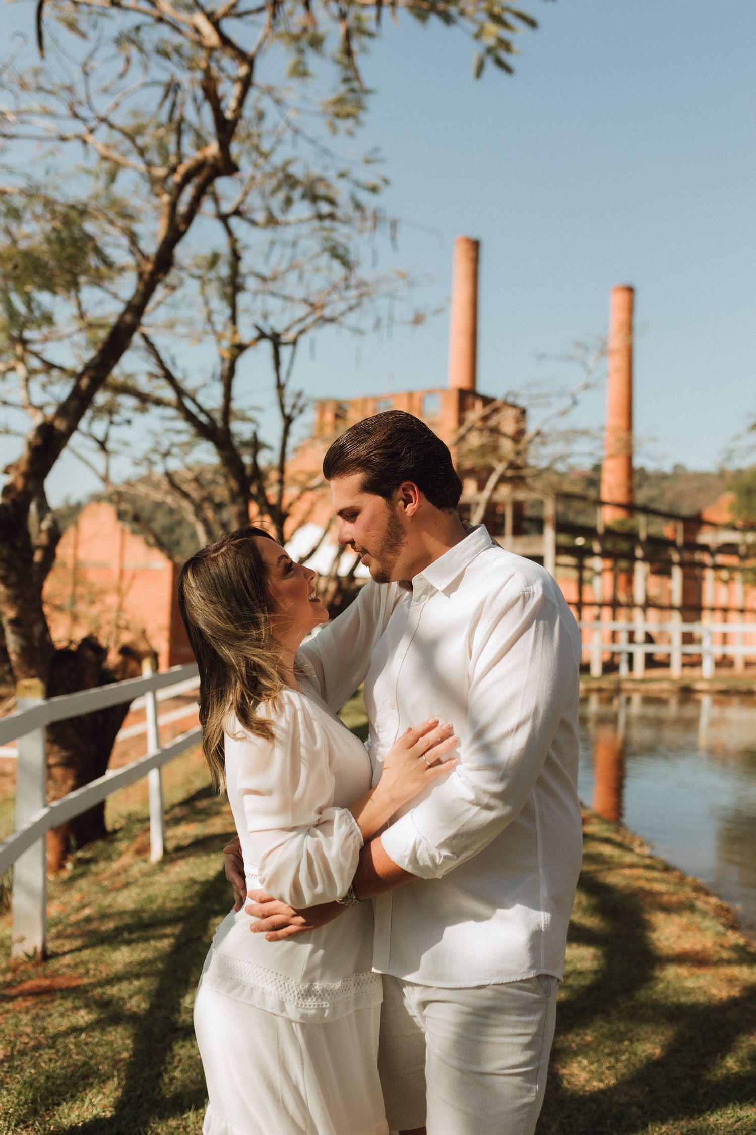 Couple in white outfits embracing near a lake with red brick industrial chimneys and trees in the background.