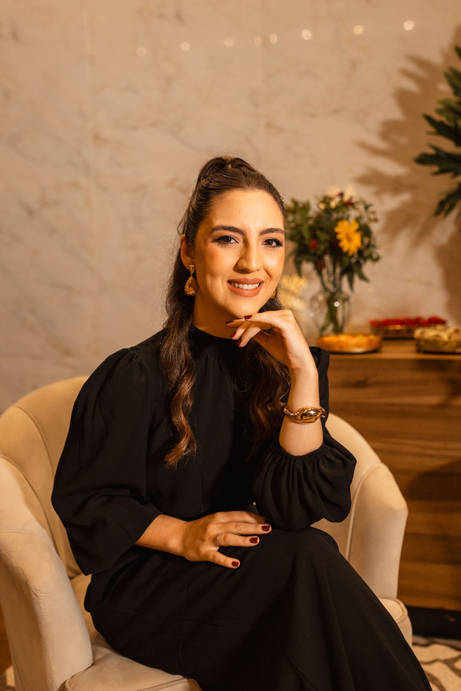 A smiling woman in a black dress sits in a beige chair, wearing gold jewelry, with flowers in the background.