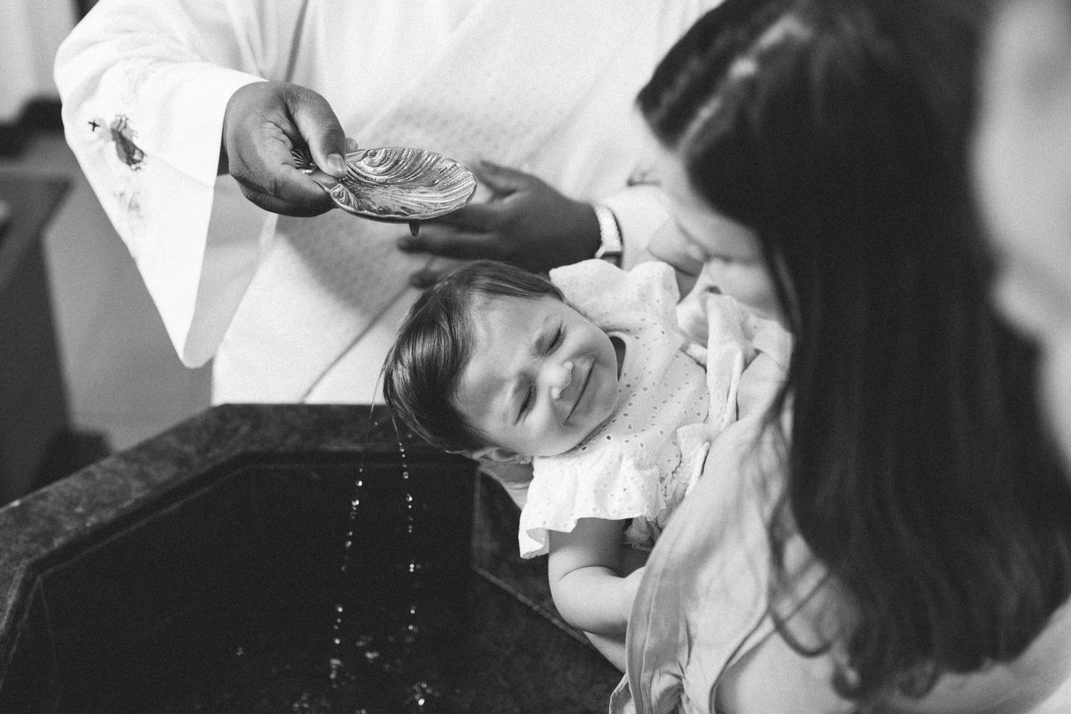 Black and white photo of a baby being baptized by a priest pouring holy water at a church font.