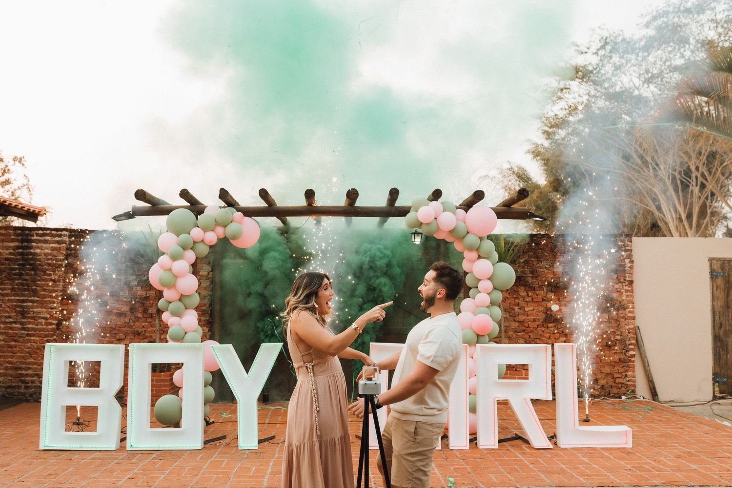Gender reveal party with couple opening box, BOY GIRL signs, balloon arch, and colorful smoke in outdoor setting.