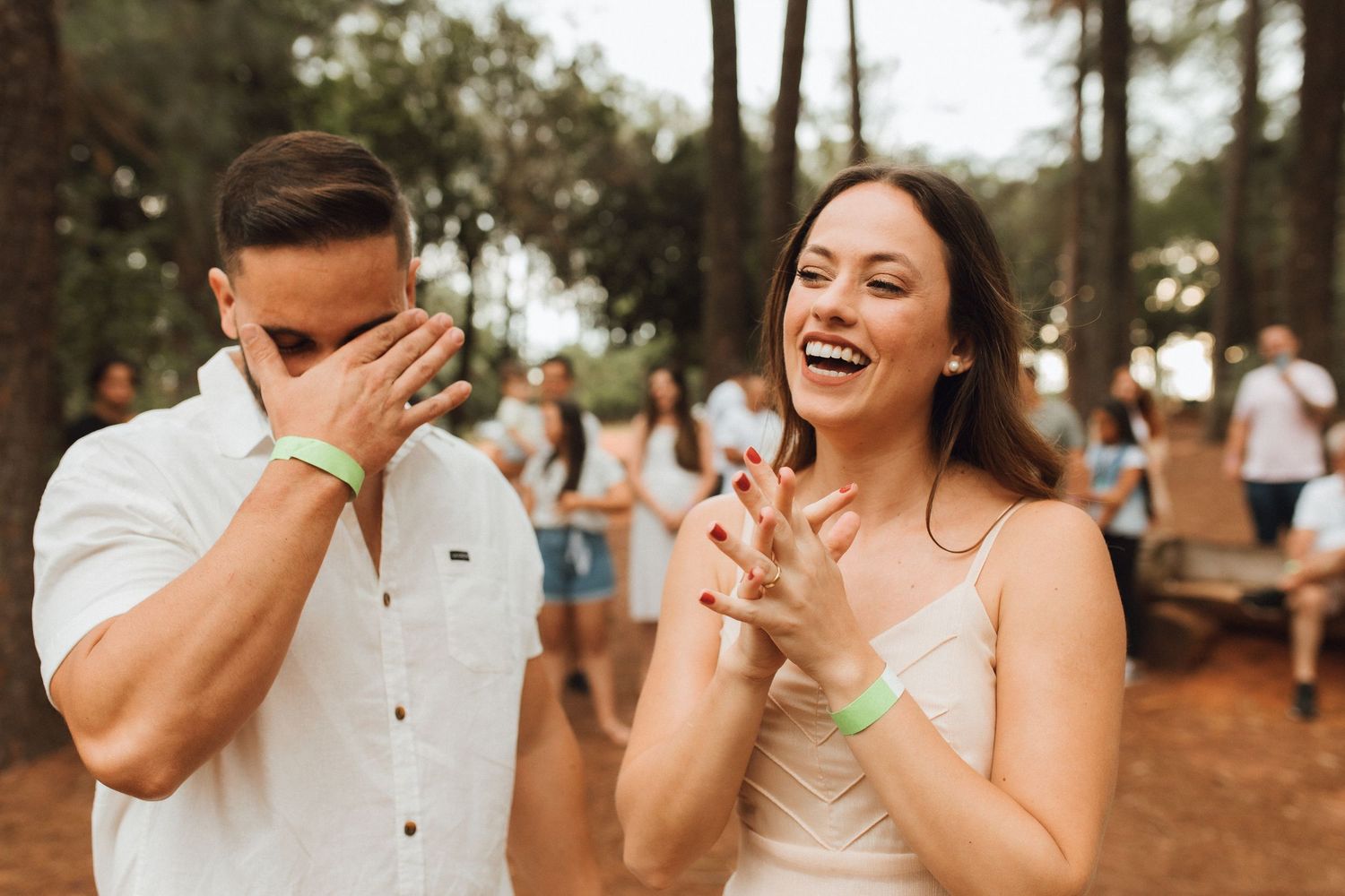 A laughing couple at an outdoor festival, woman clapping and man covering his face, both wearing green wristbands.
