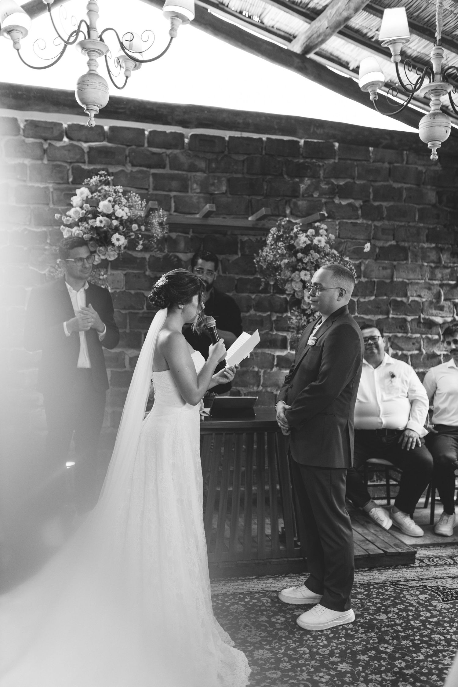 Bride reads wedding vows to groom at rustic brick outdoor ceremony with floral arrangements.