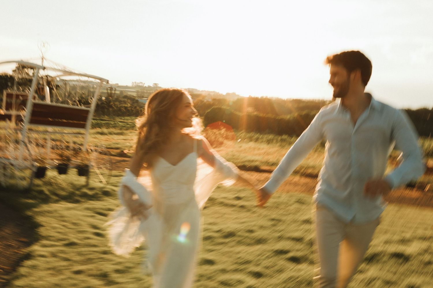 Couple in white holding hands running through golden sunlit field during romantic outdoor engagement session.