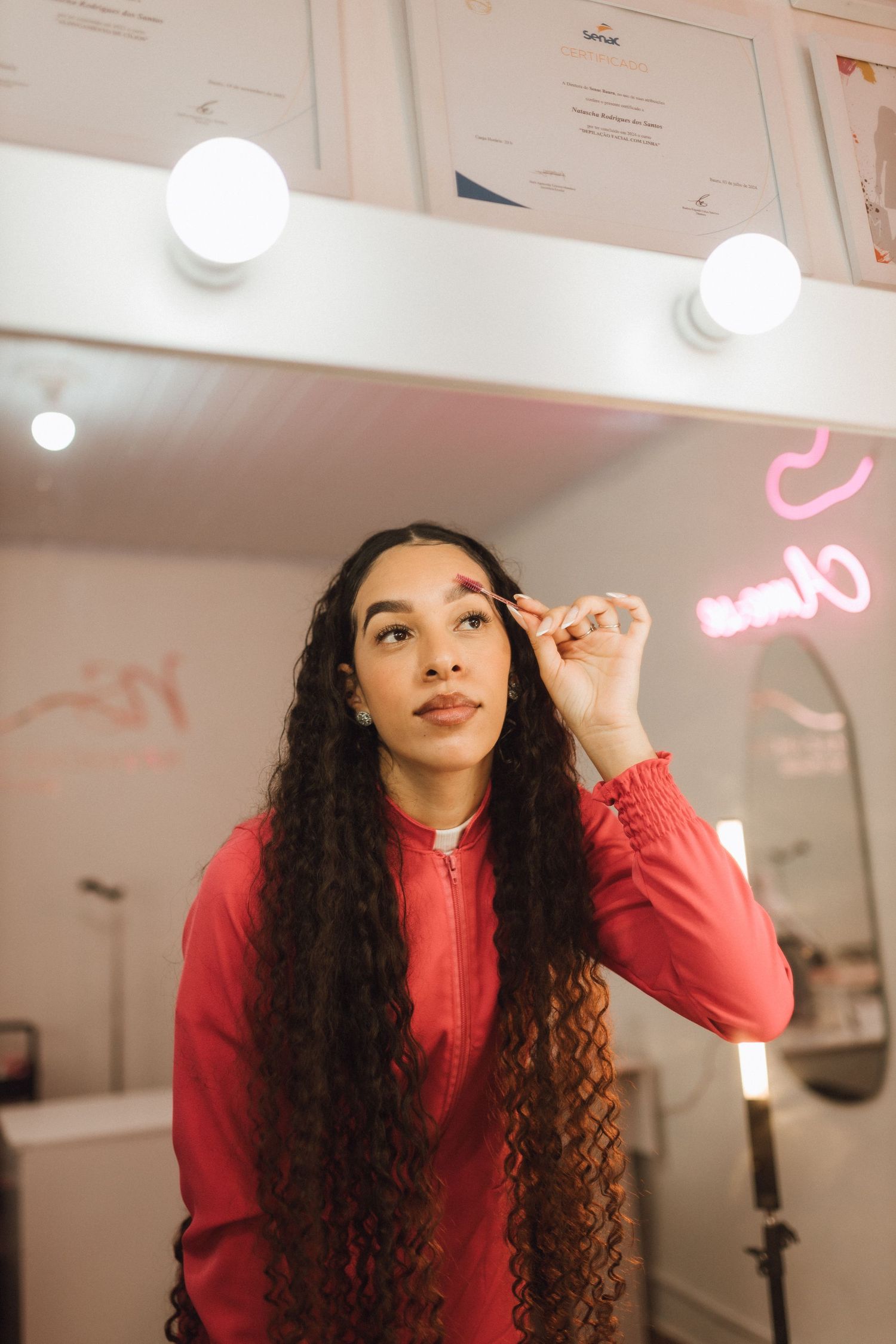Young woman with curly hair grooming eyebrows in illuminated vanity mirror salon.