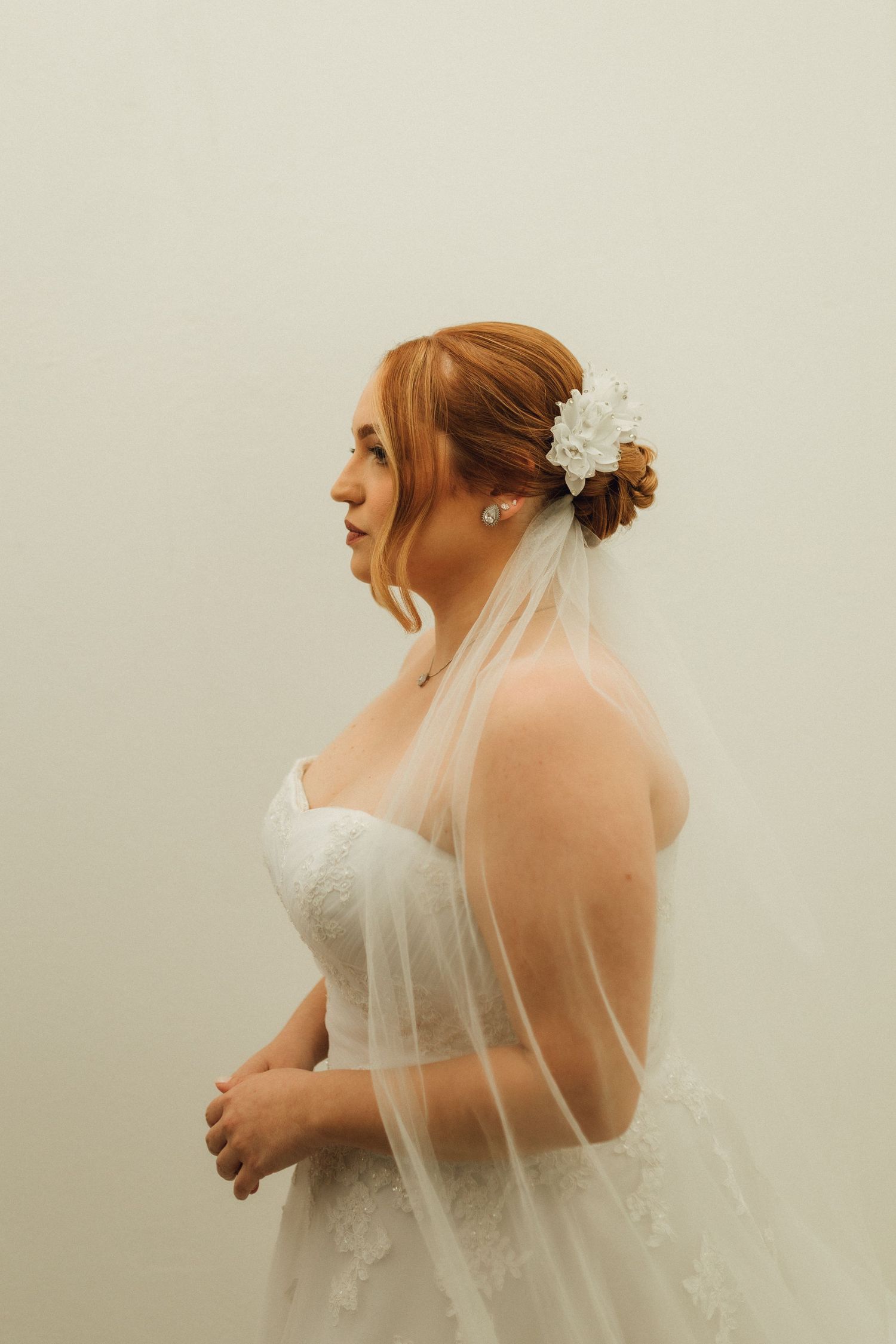 Side profile of redhead bride in strapless lace gown with white floral hair accessory and veil.