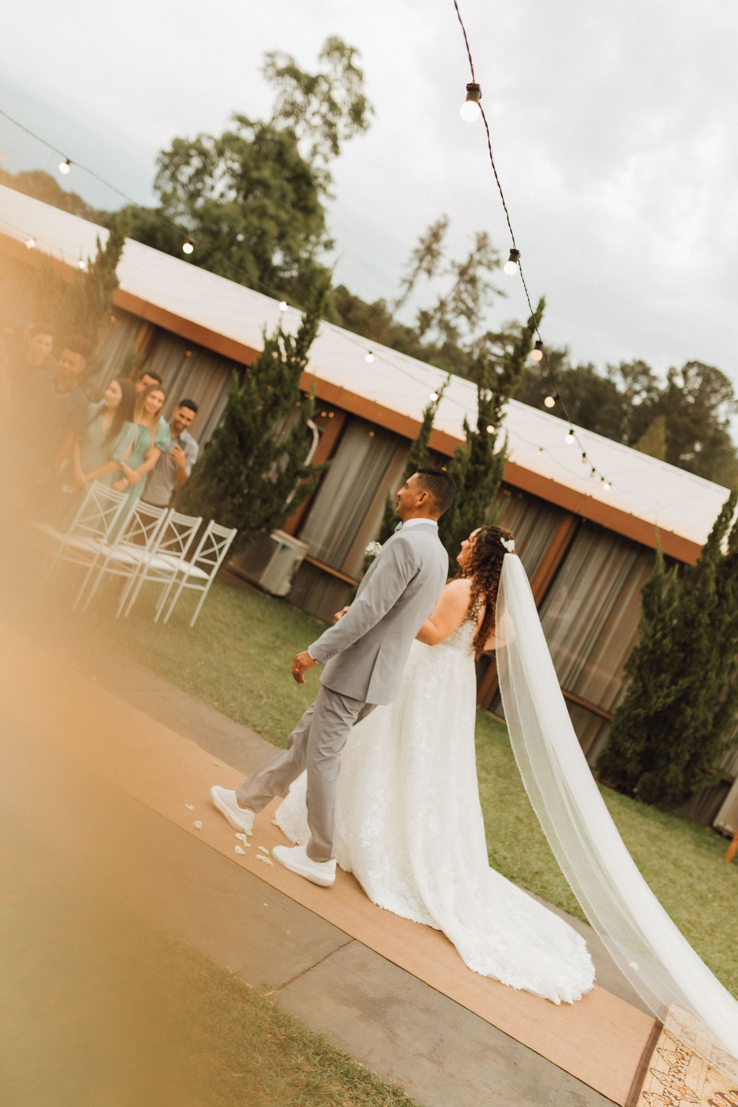 Bride and groom walk down outdoor wedding aisle with string lights and cypress trees in background.