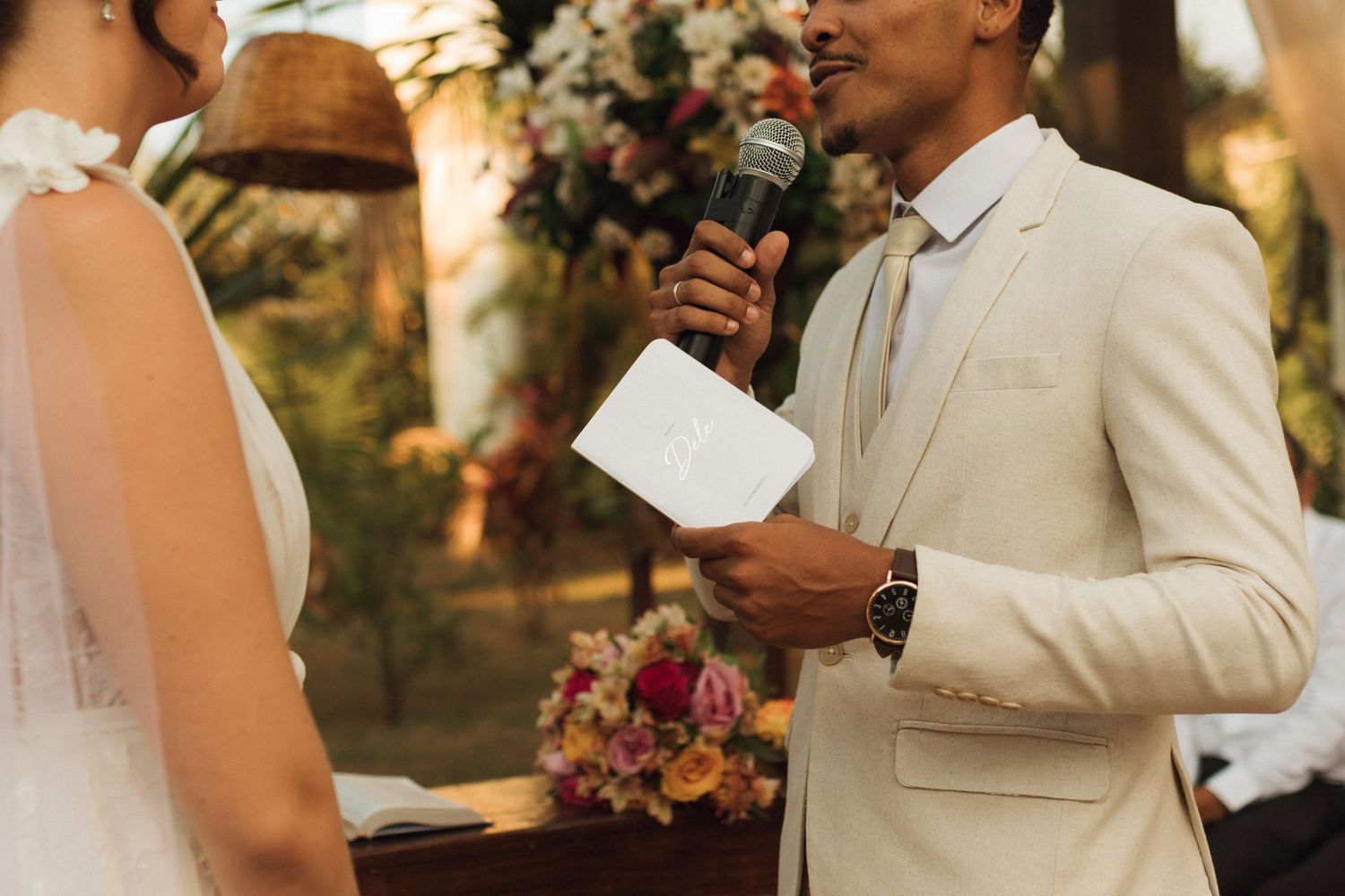 Groom reading wedding vows with a microphone at an outdoor ceremony with floral decor.