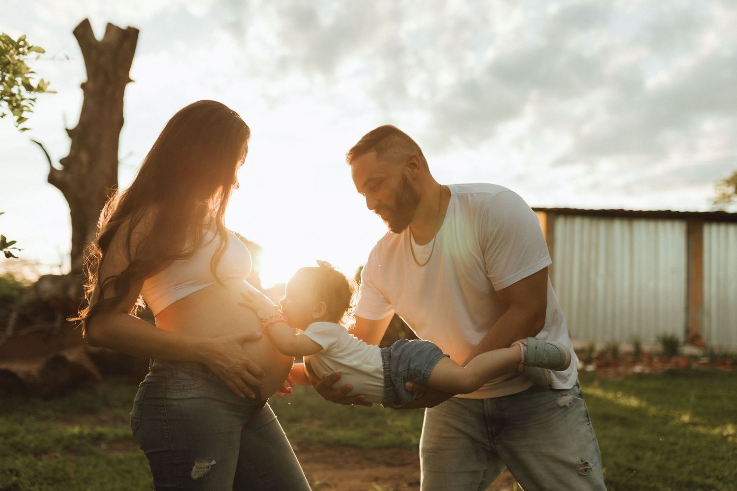 A couple shares a joyful moment with their baby outdoors at golden hour, bathed in warm sunset light.