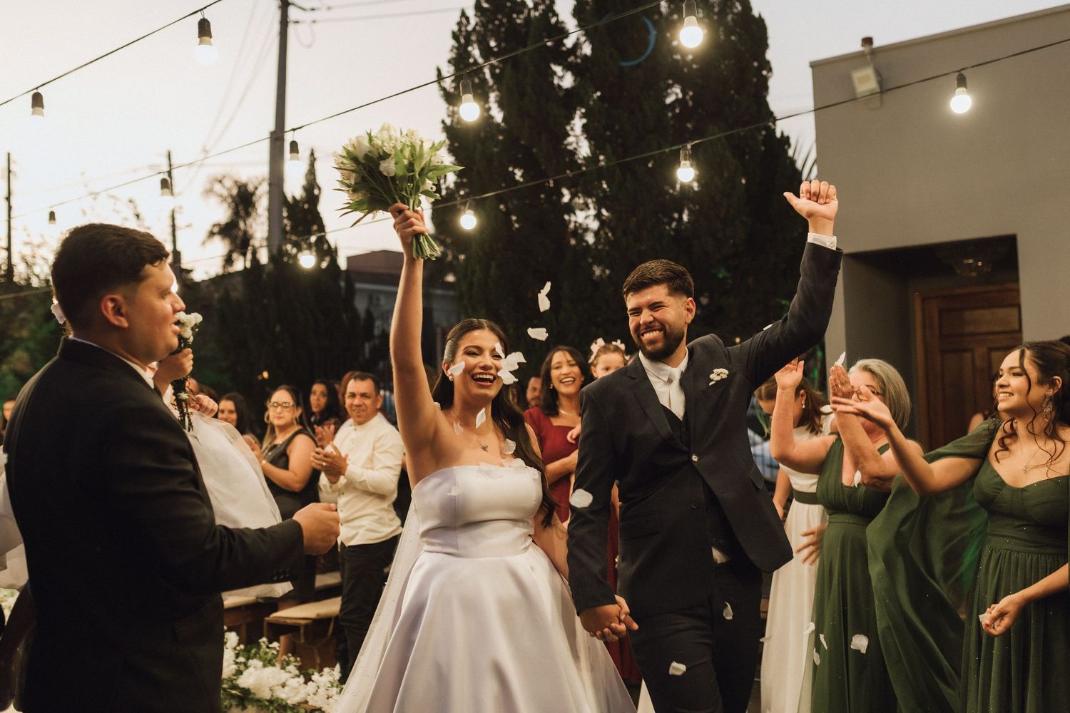 Joyful bride and groom exit wedding ceremony with sparklers, guests in green dresses cheering outdoors.
