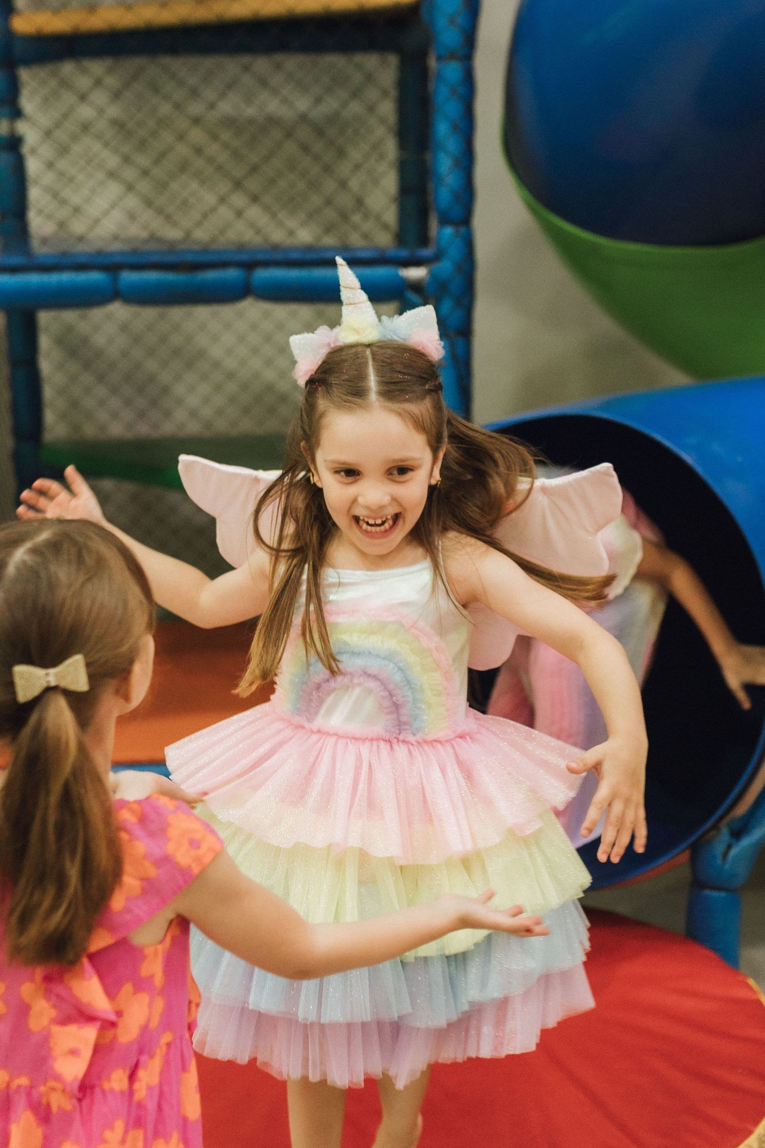 Happy girl in unicorn costume with rainbow dress and wings playing at a colorful indoor birthday party.