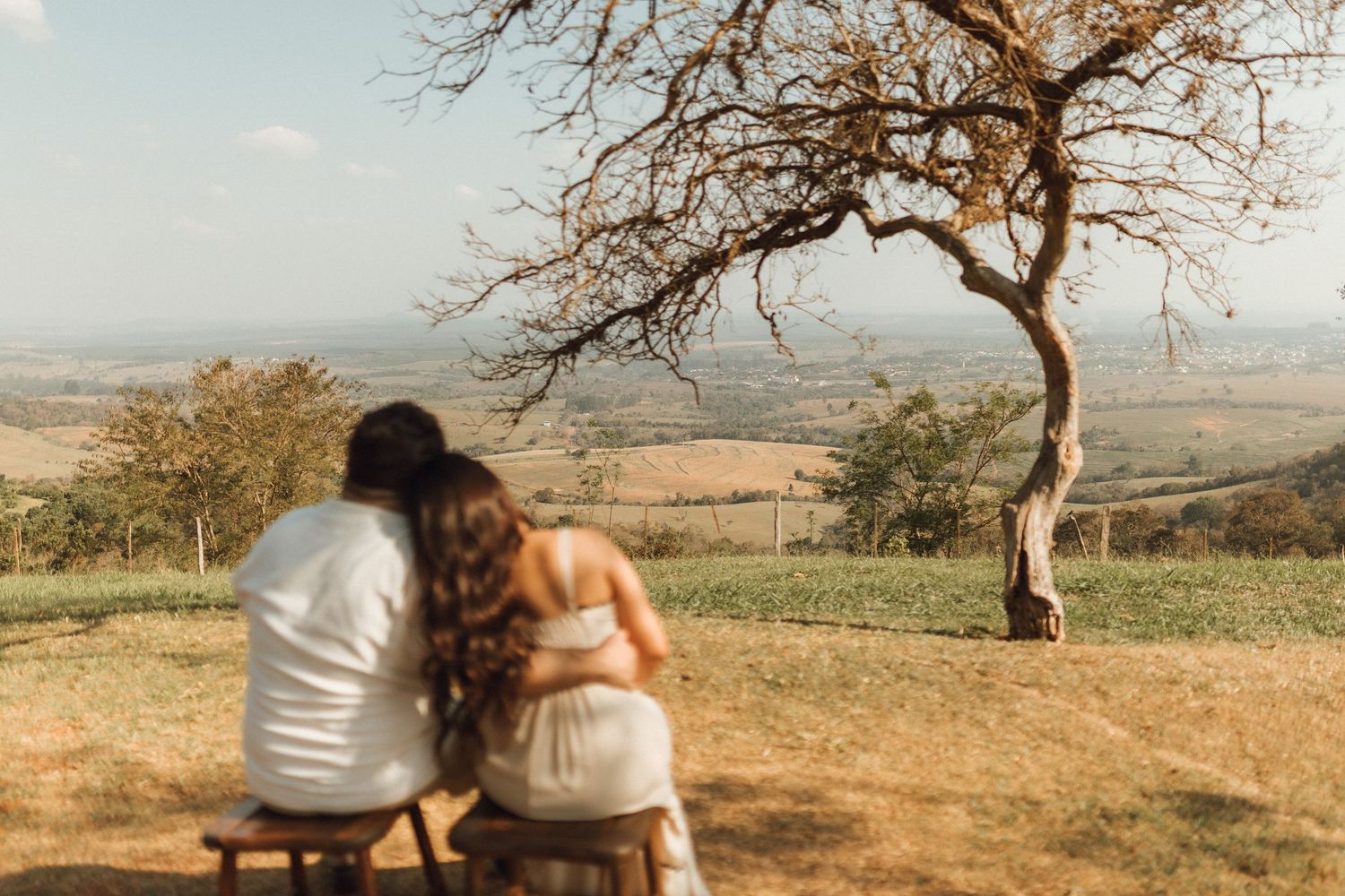 Couple sitting on a bench embracing, overlooking rolling countryside landscape with a bare tree nearby.