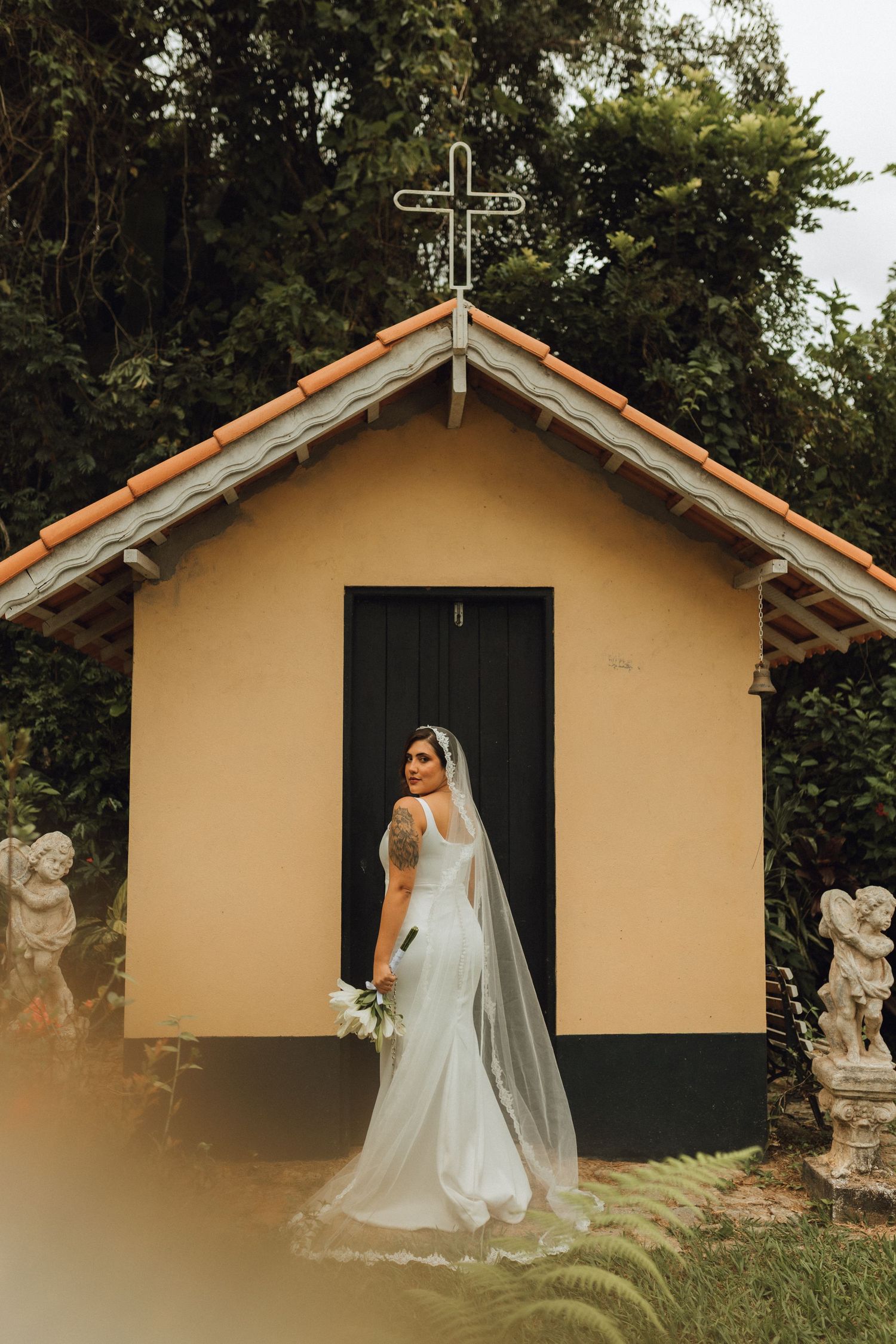 Bride in white gown and veil posing before a small yellow chapel with a cross.